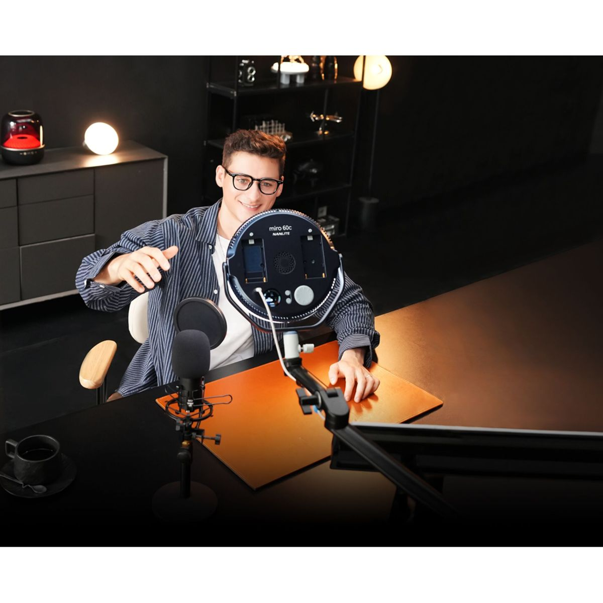 Ein Mann mit Brille hält ein Ringlicht und lächelt. Ein Mikrofon ist vor ihm. Eine Kaffeetasse steht auf dem Tisch.