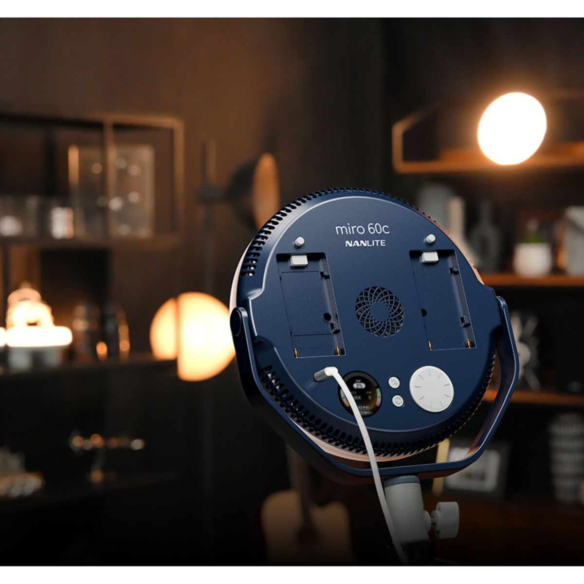 Eine dunkelblaue Nanlite Miro 60C Leuchte steht auf einem Ständer, mit einem weißen Kabel. Warme Glühbirnen im Hintergrund.