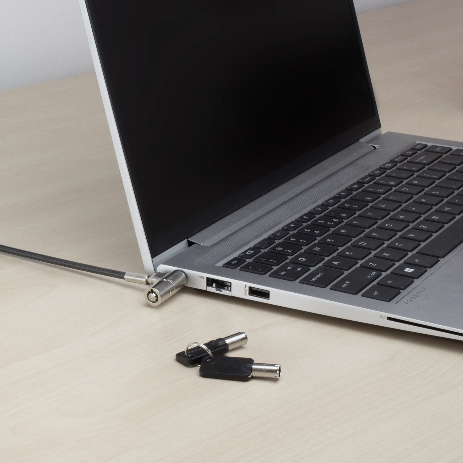 Laptop mit Kabelschloss gesichert. Schlüssel liegen neben dem Laptop auf einem Holztisch. Der Bildschirm ist schwarz, die Tastatur sichtbar.