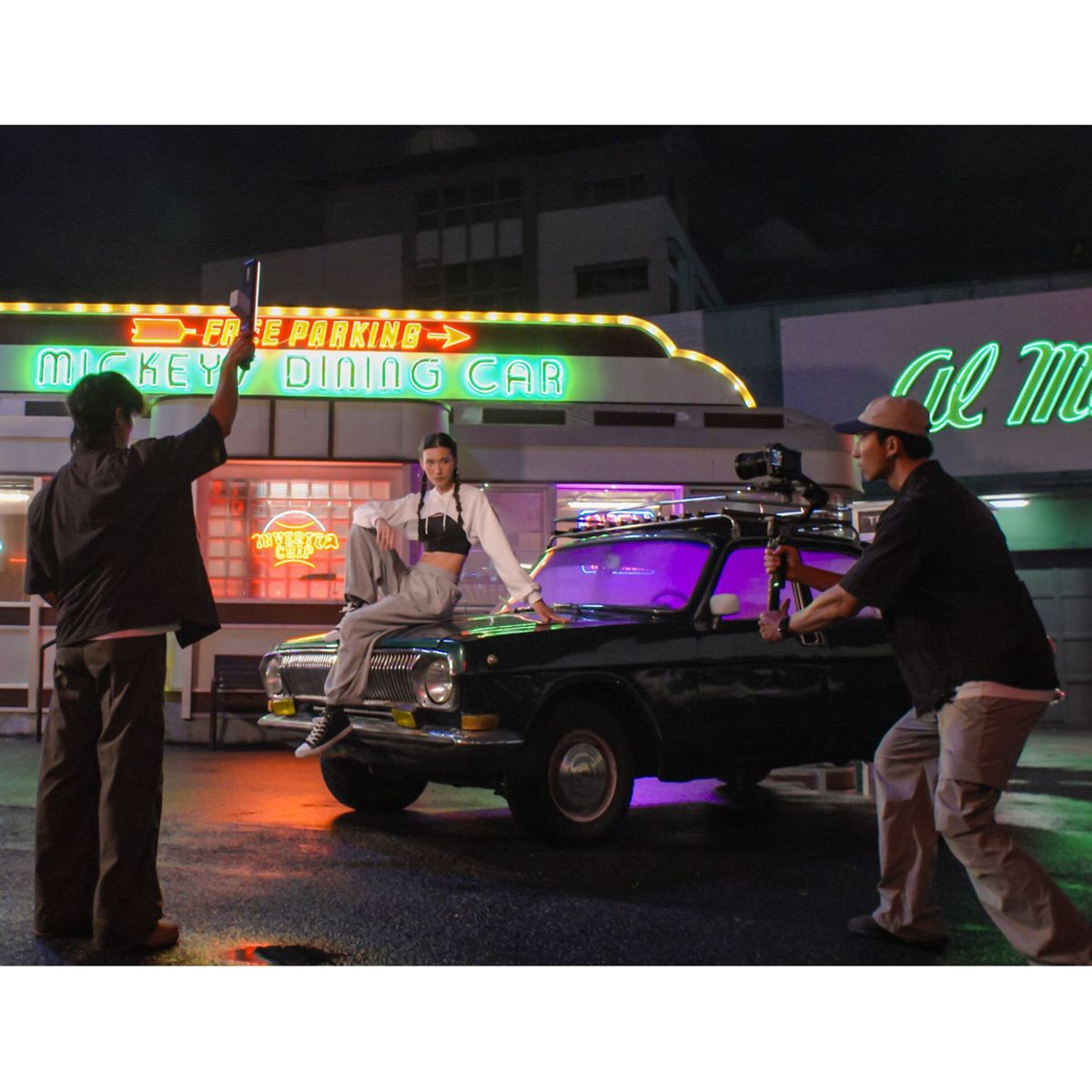Una donna seduta su un'auto. Due uomini la filmano. Insegne al neon sullo sfondo, tra cui 'Mickey Dining Car'.