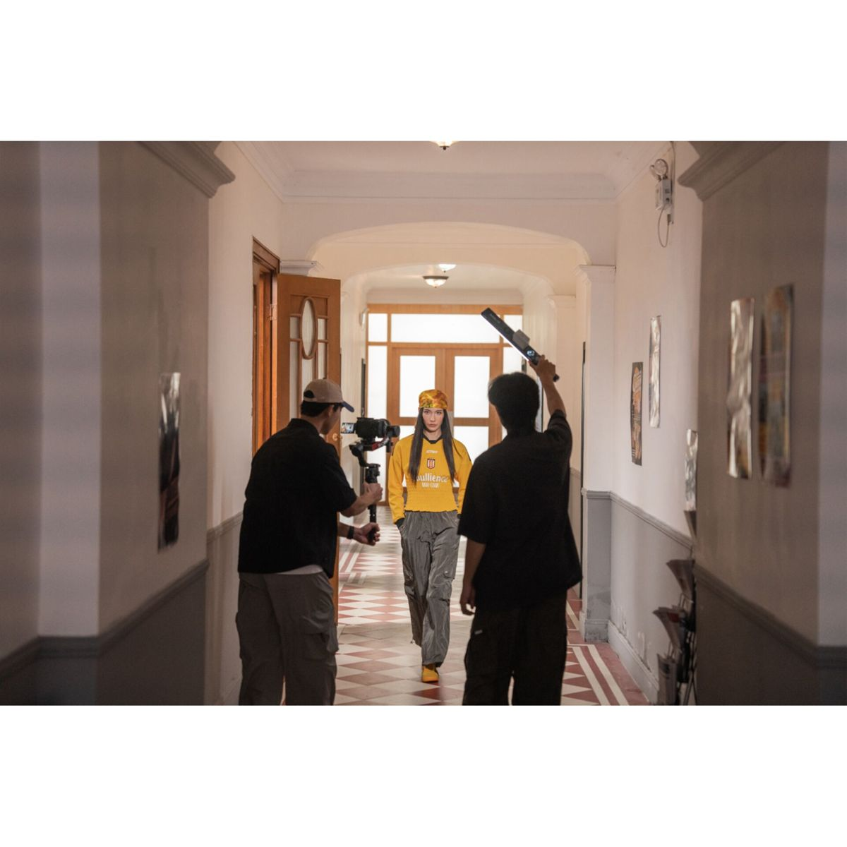 Una donna cammina in un corridoio. Due persone la filmano. Uno tiene una luce, l'altro una telecamera. La donna indossa giallo e grigio.
