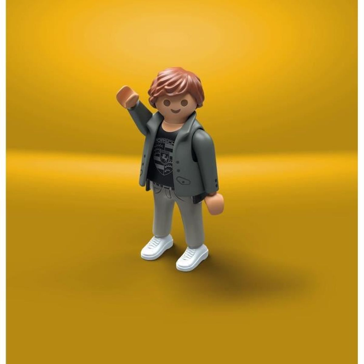 Una figura de Playmobil con cabello castaño y una chaqueta está saludando. Lleva una camiseta, pantalones grises y zapatos blancos. El fondo es amarillo.