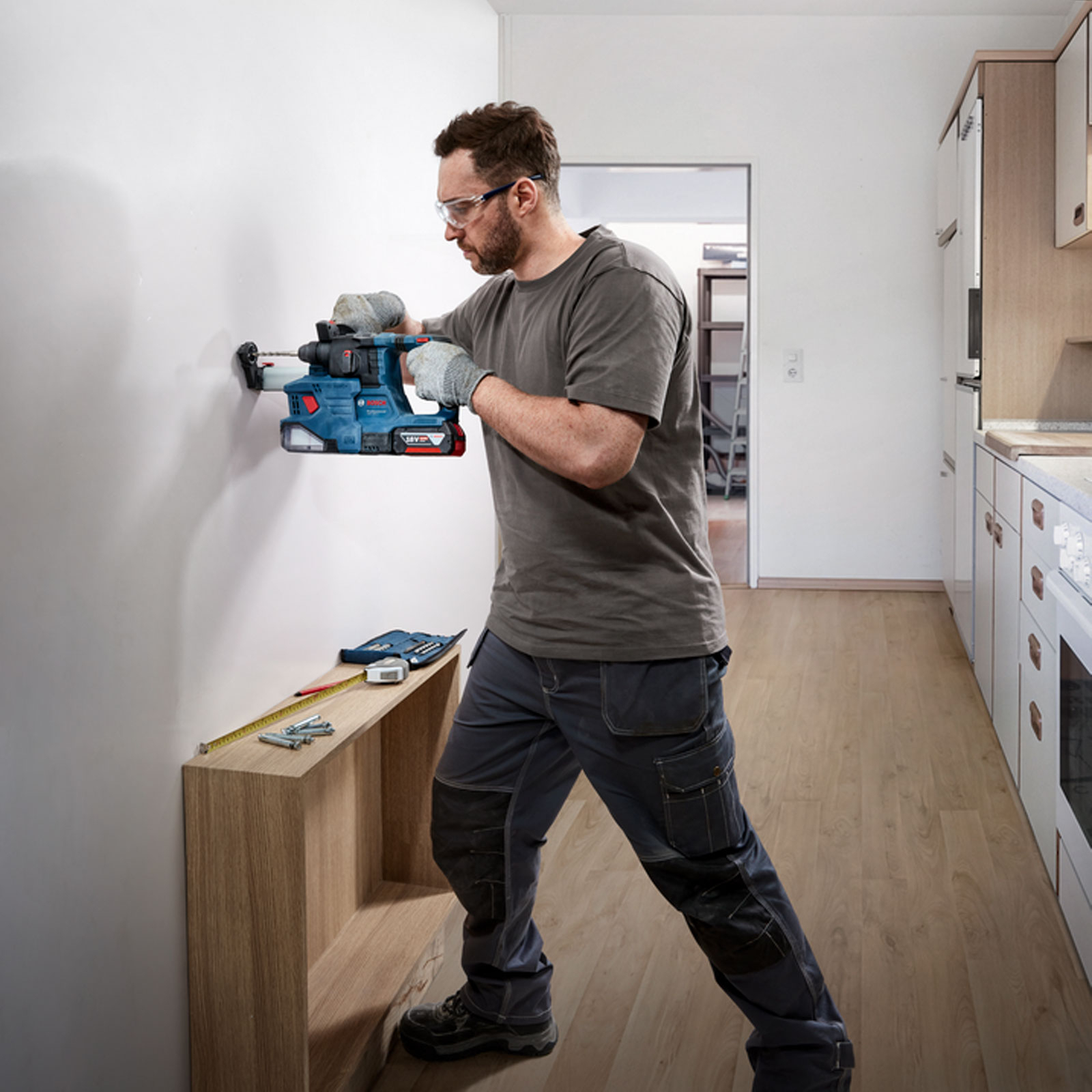 Ein Mann bohrt mit einem Elektrowerkzeug in eine Wand. Darunter ein Regal mit Werkzeugen. Küche im Hintergrund sichtbar.