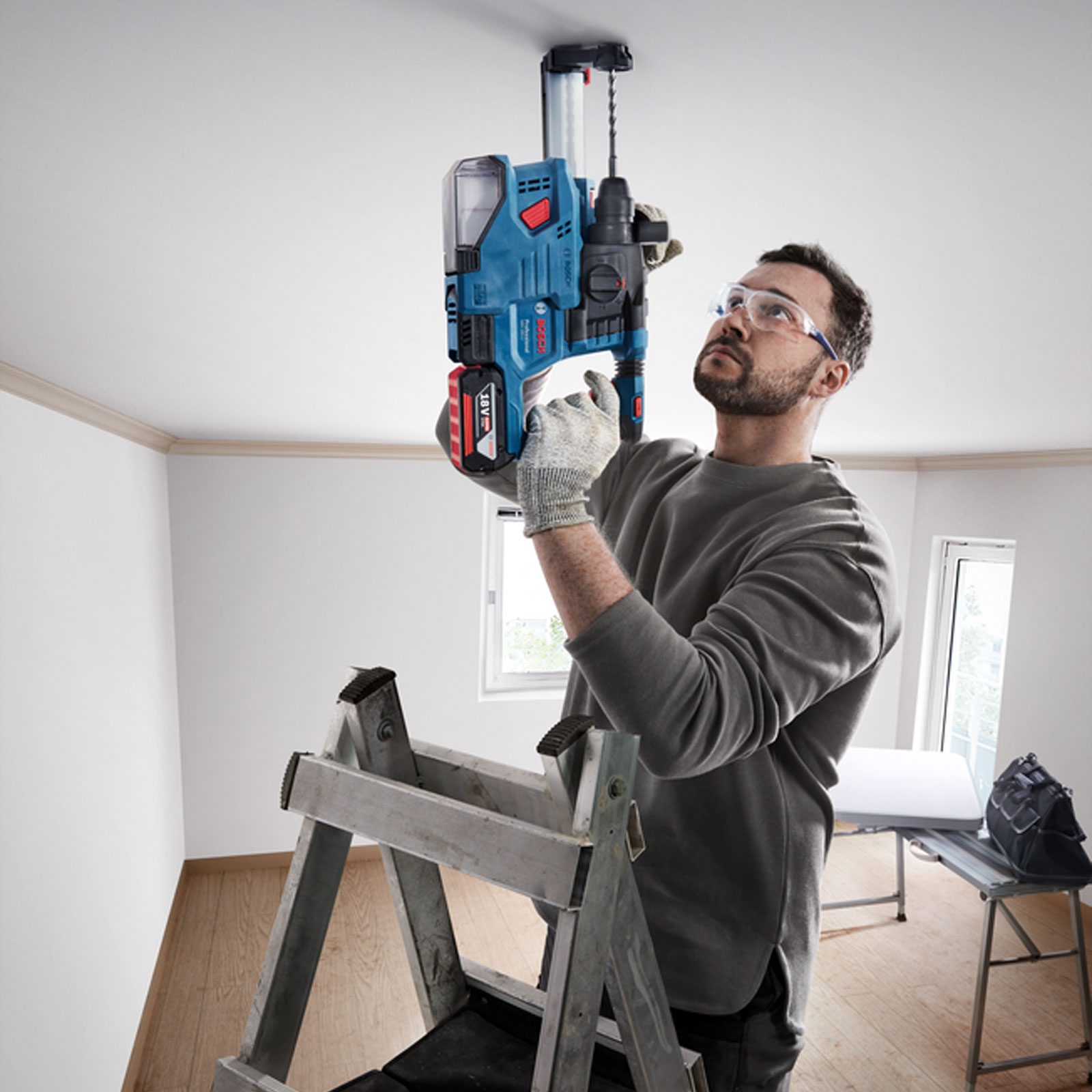 Ein Mann benutzt eine Bohrmaschine an der Decke und steht auf einer Leiter. Er trägt Schutzbrille und Handschuhe, die Bohrmaschine ist blau und schwarz.