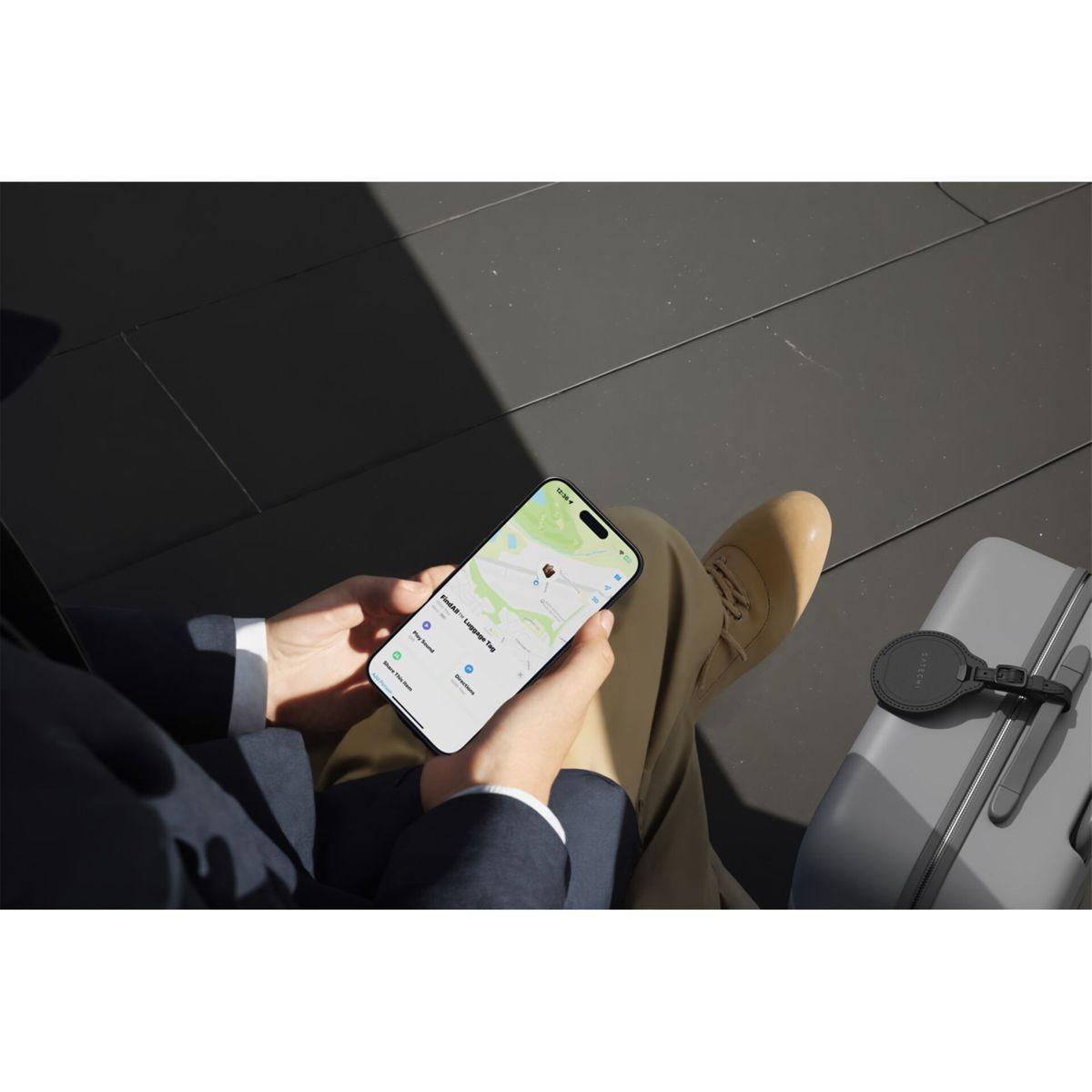 Eine Person hält ein Telefon mit einer Karte, daneben ein Gepäckanhänger und ein Koffer. Die Person trägt einen Anzug und beige Schuhe.