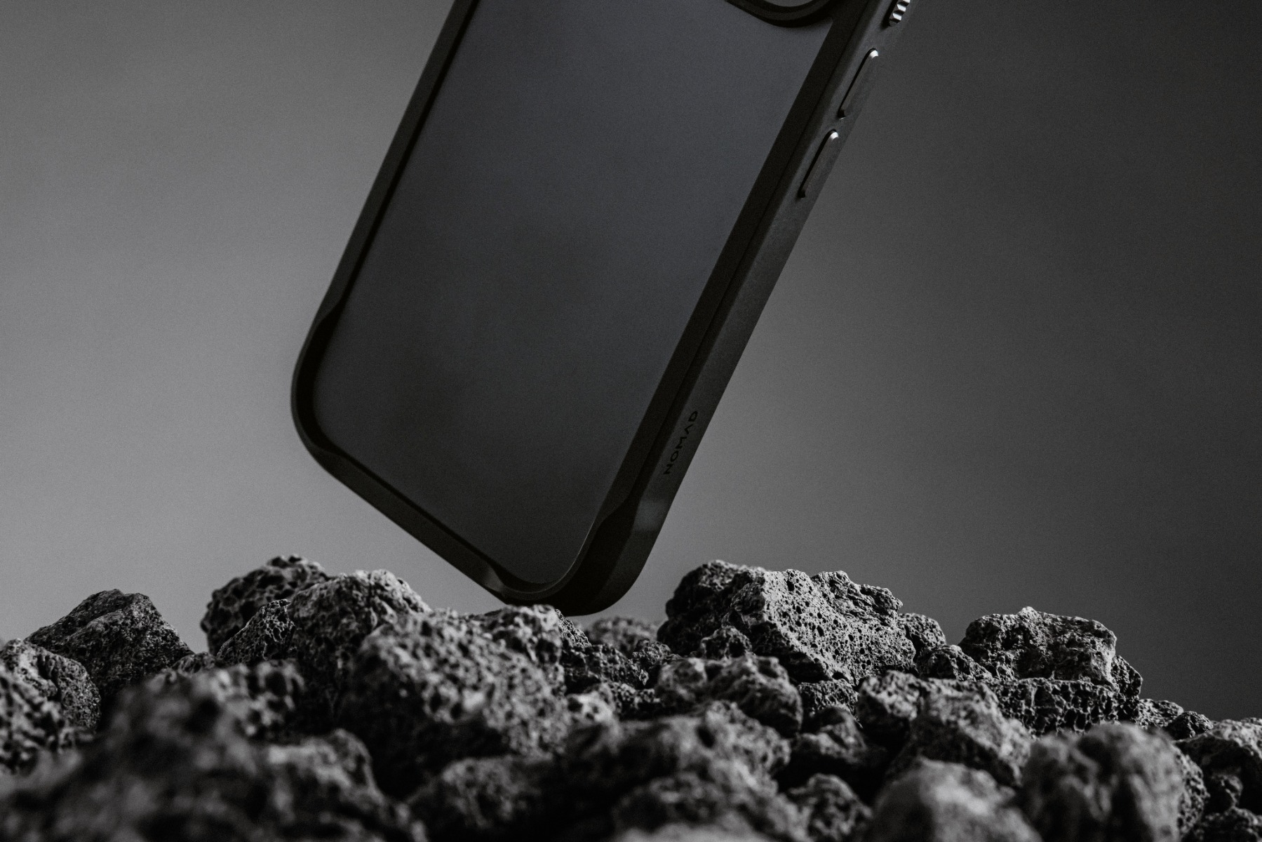 Custodia nera per telefono sospesa su rocce scure e strutturate. La custodia ha bordi arrotondati. Lo sfondo è un grigio sfumato.