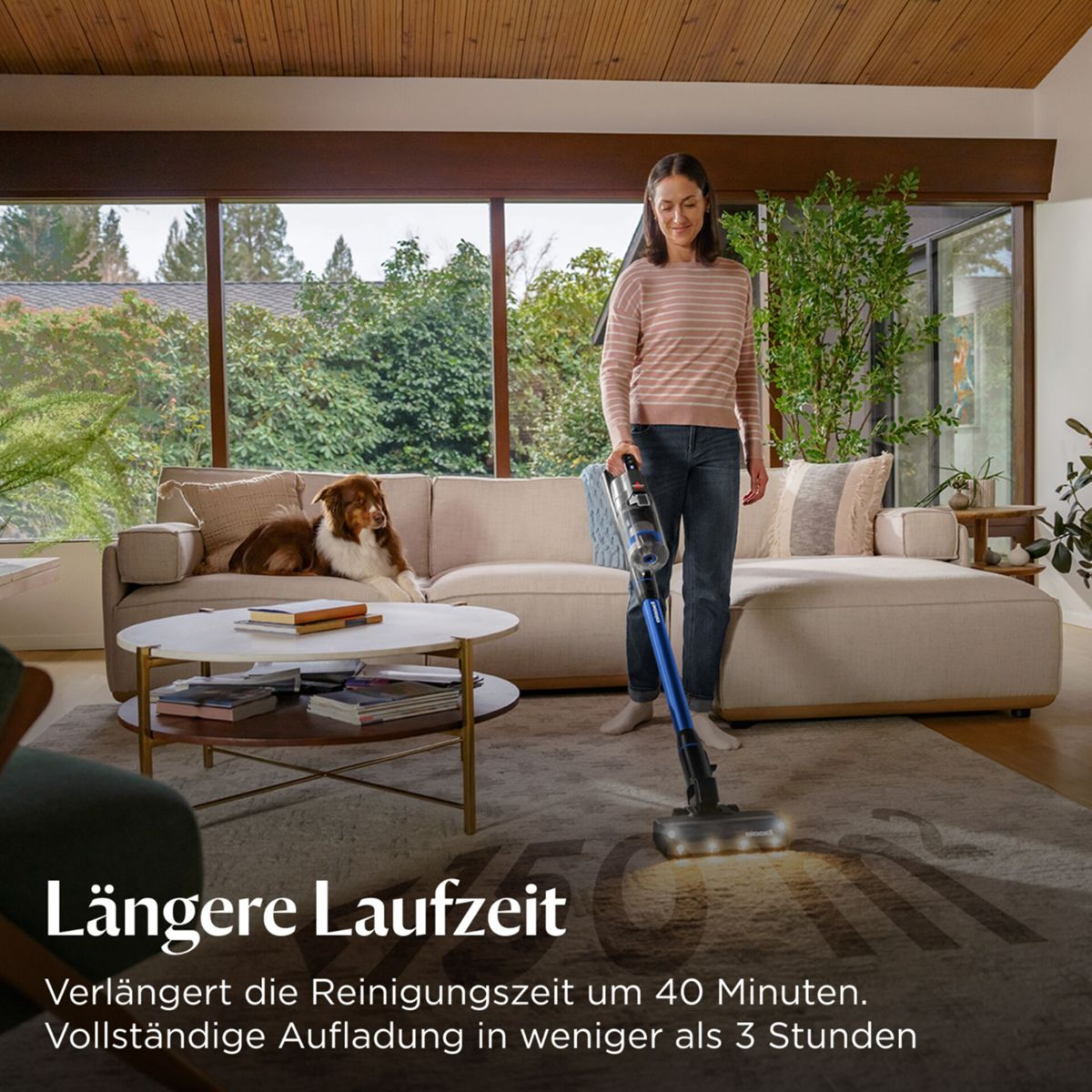 Eine Frau saugt einen Teppich in einem Wohnzimmer. Ein Hund liegt auf einem Sofa. Der Raum hat große Fenster und eine Holzdecke.