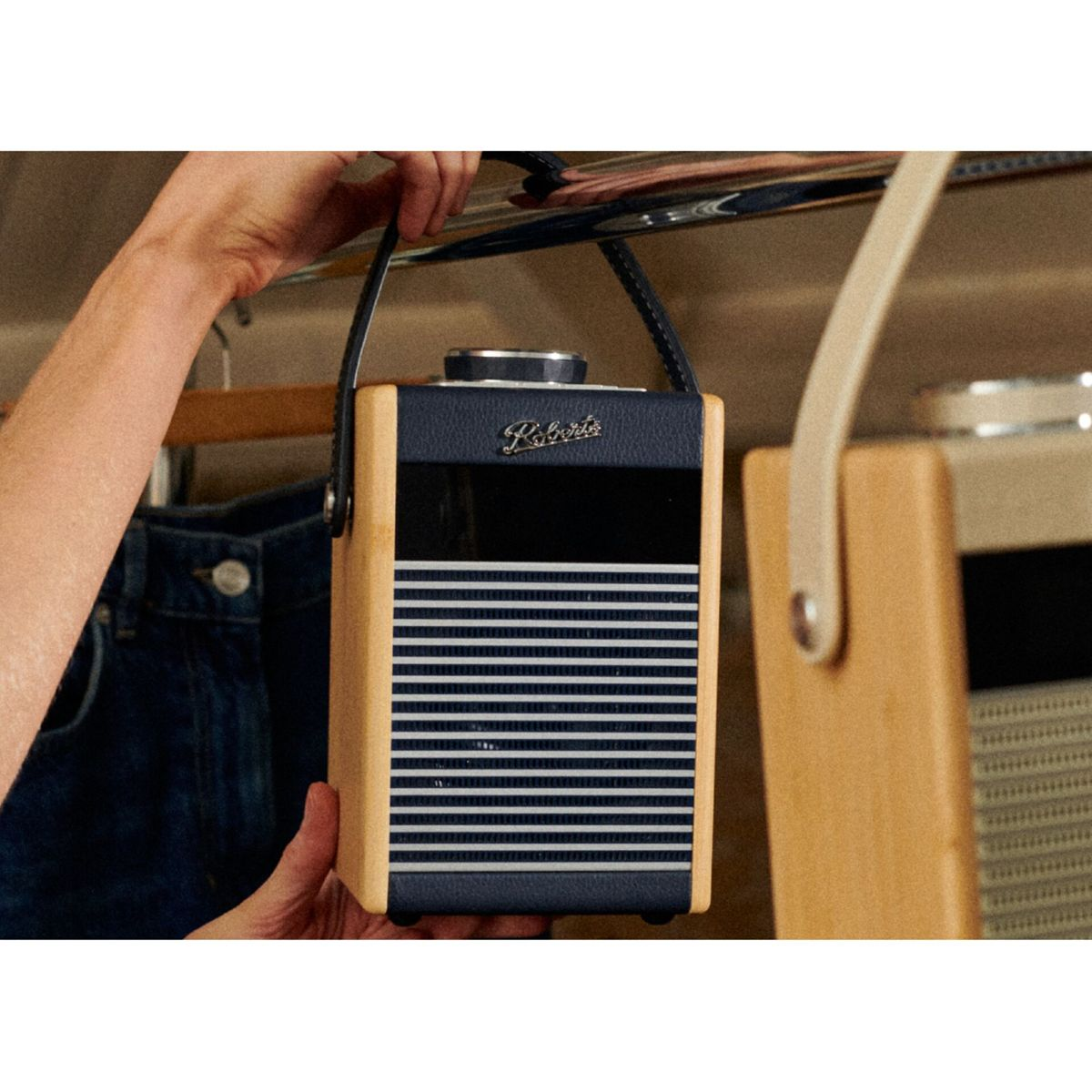 Una persona tiene in mano una radio Roberts blu navy e beige. La radio ha una maniglia e lo sfondo mostra un armadio con vestiti.