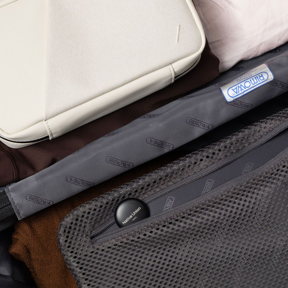 Kofferinnenraum mit weißer Tasche, Kleidung und einem schwarzen Native Union Gerät in einer Netztasche. Rimowa-Marke vorhanden.