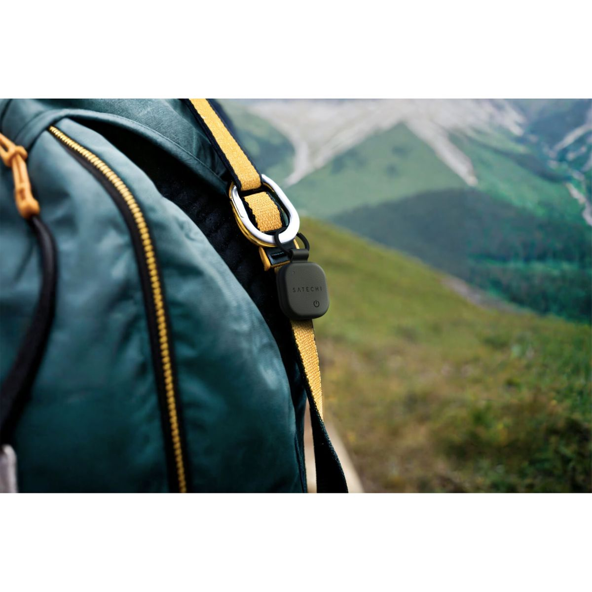 Ein grüner Rucksack mit gelbem Gurt und einem schwarzen Satechi-Gerät. Berge sind im Hintergrund sichtbar.