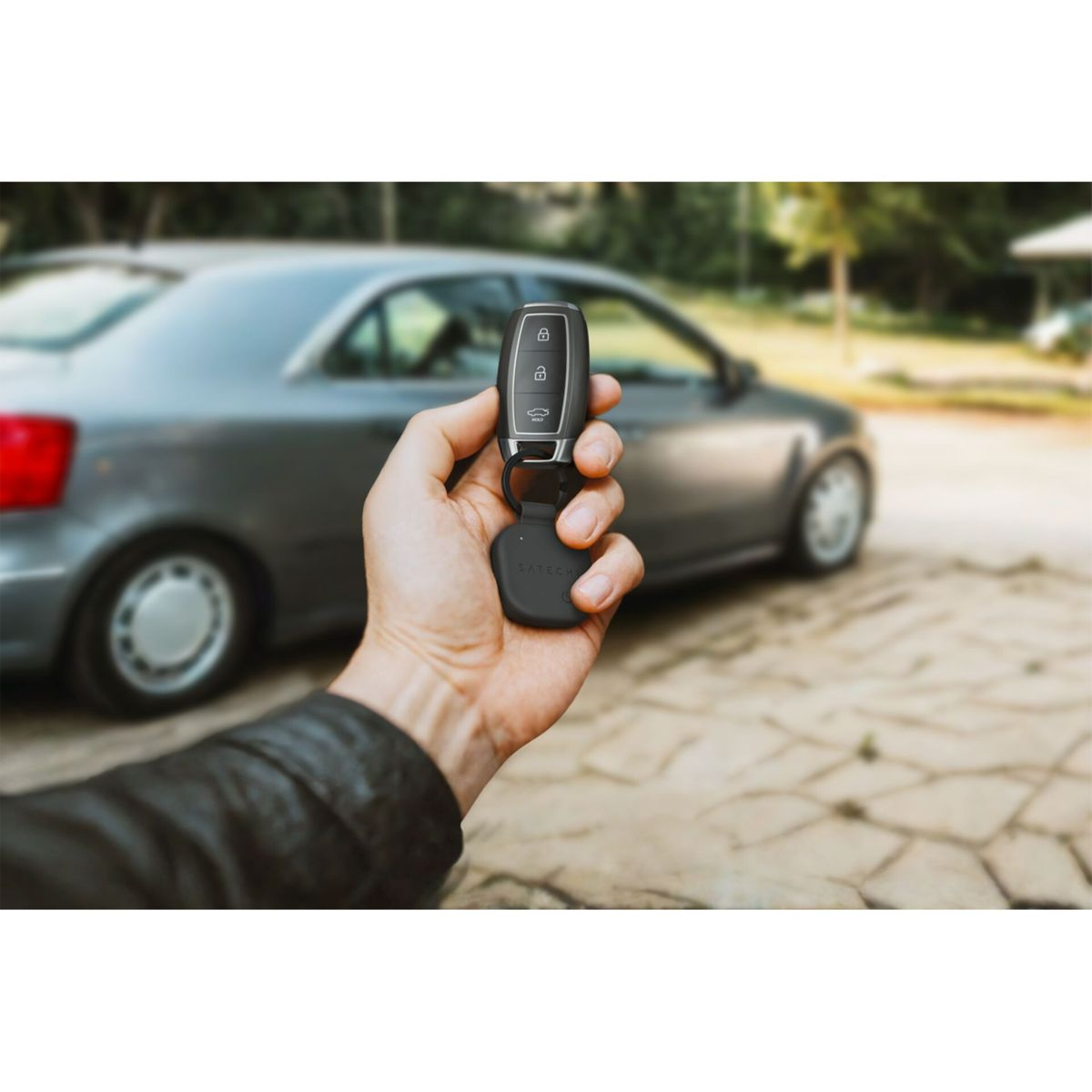 Eine Hand hält einen Autoschlüsselanhänger vor einem grauen Auto. Der Schlüssel hat Schloss-, Entriegelungs- und Kofferraumtasten. Der Hintergrund ist unscharf.