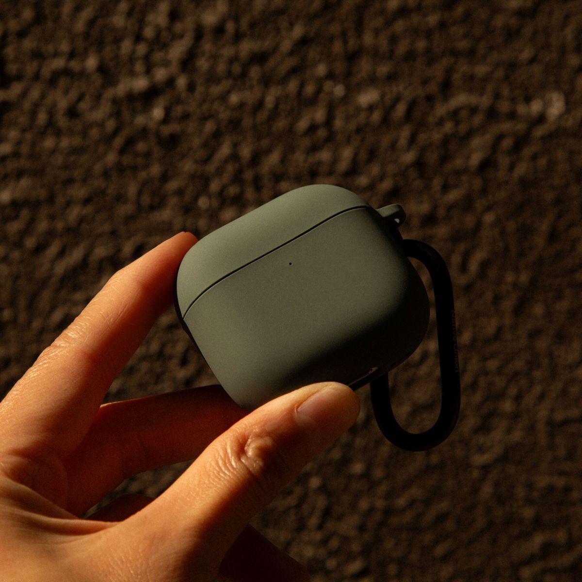 Eine Hand hält ein grünes AirPods-Etui mit schwarzem Karabiner vor einem strukturierten, braunen Hintergrund. Sonnenlicht erzeugt Schatten.