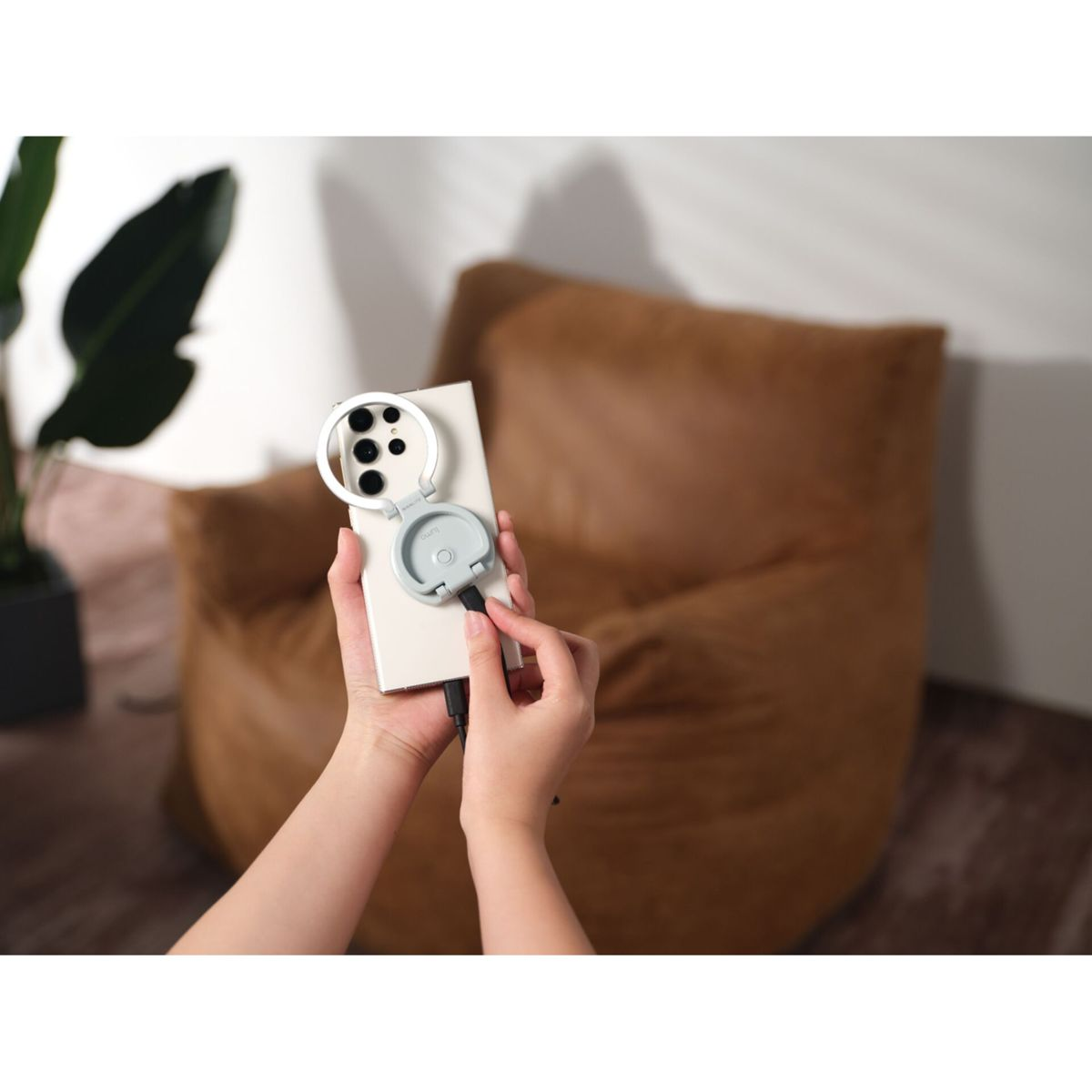 Hände halten ein Smartphone mit Ringlichtaufsatz. Im Hintergrund ist ein brauner Sitzsack zu sehen.