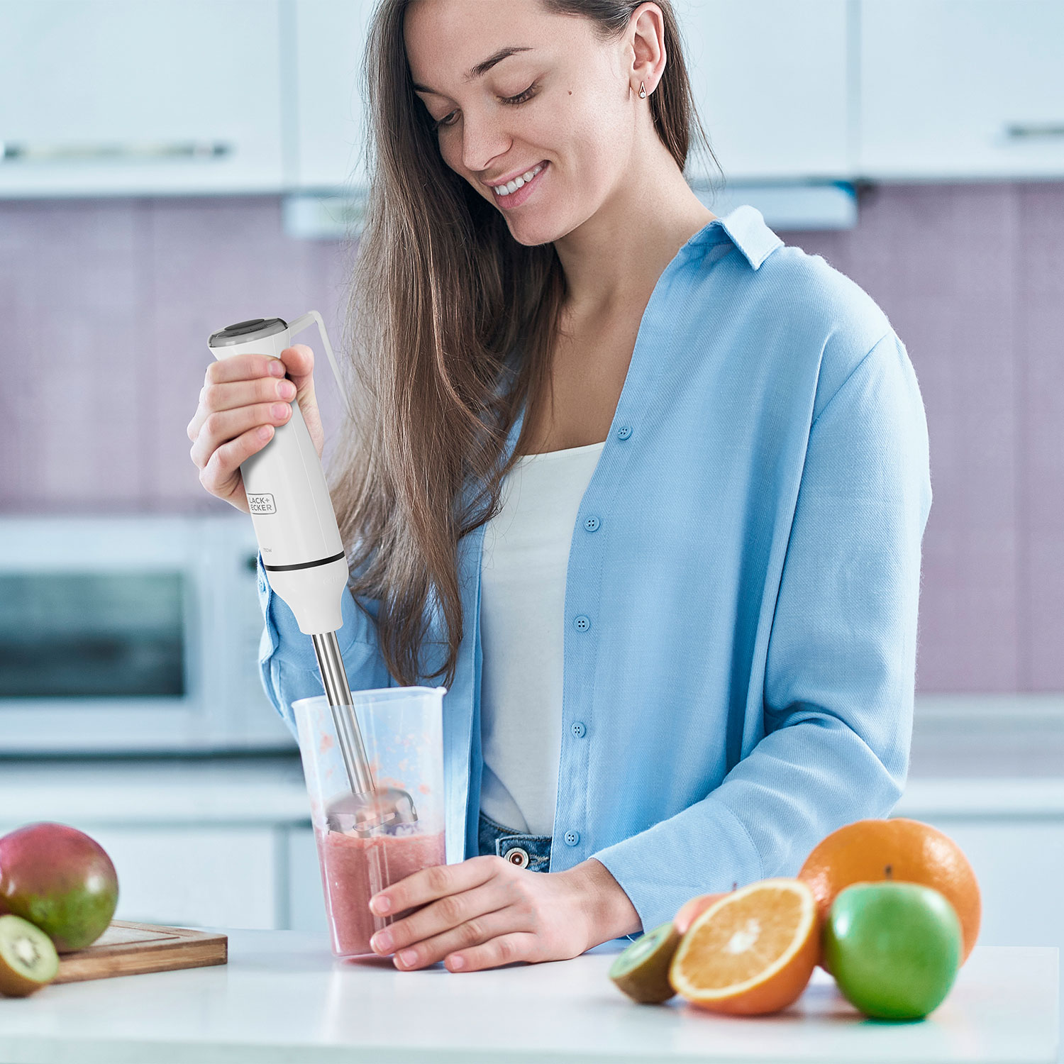 Una donna sorride mentre frulla un frullato rosa con un frullatore a immersione. Frutta come arance, mele e kiwi sono sul tavolo.