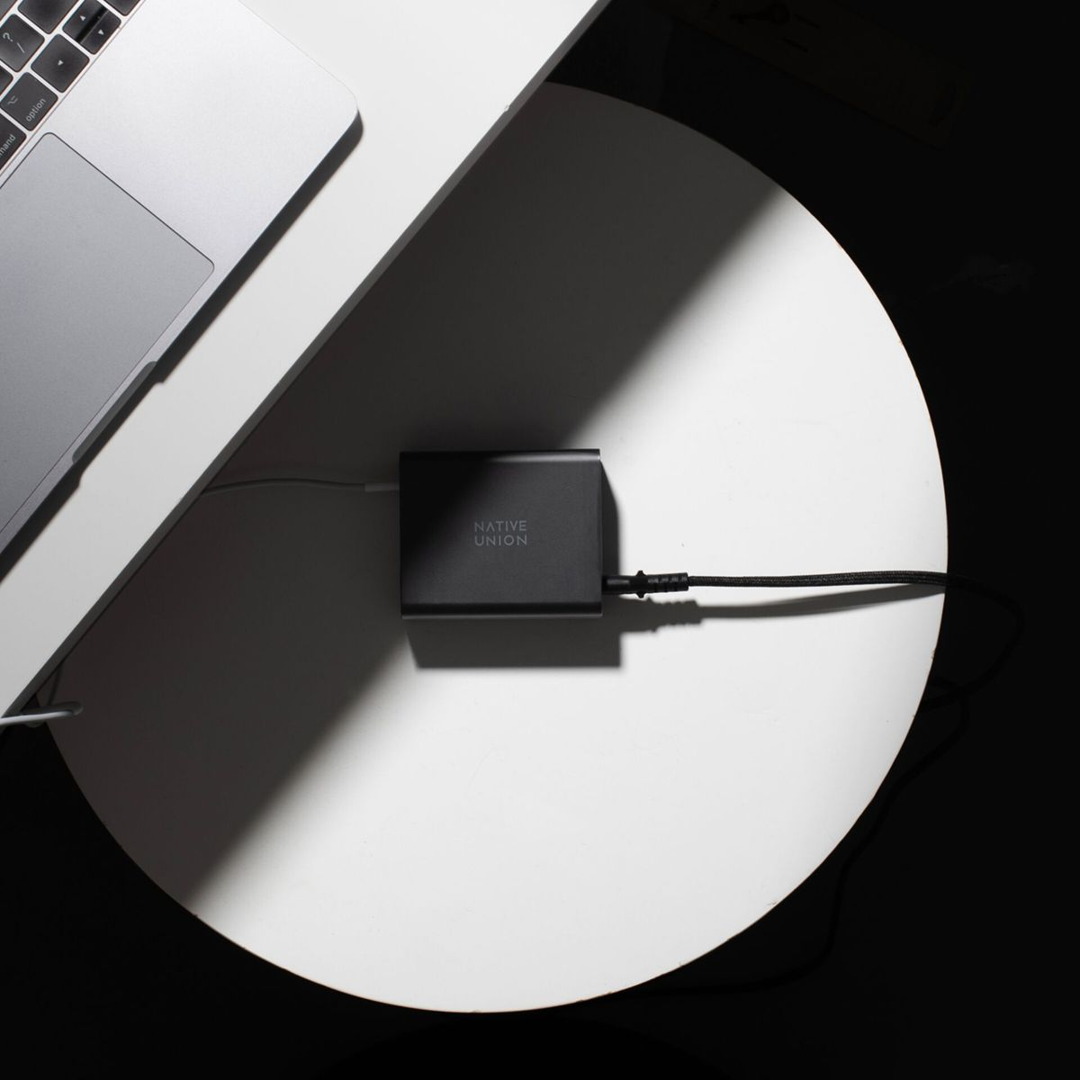 Un ordinateur portable et un adaptateur secteur Native Union noir sont sur une table blanche. Les câbles relient les appareils. L'ombre crée un contraste.