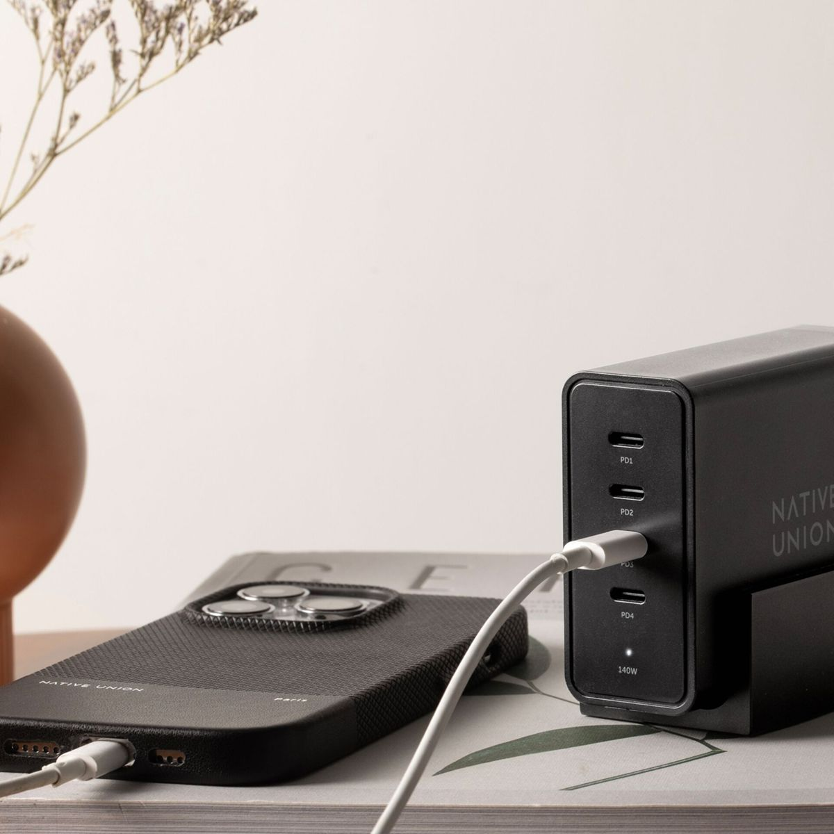 Téléphone noir avec un câble blanc en charge, à côté d'un chargeur noir. Un livre et un vase avec des plantes sont également visibles.