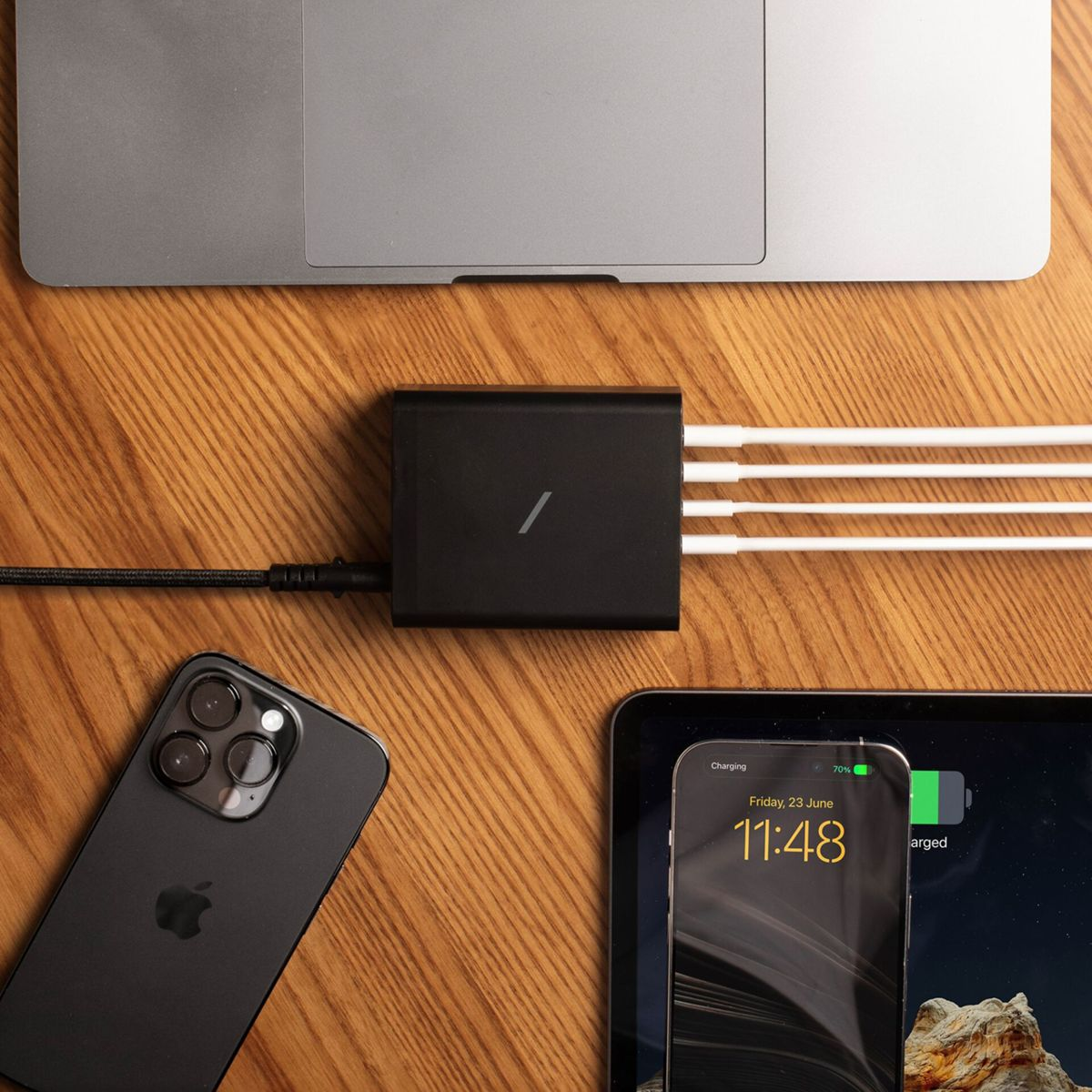 Un hub noir avec des câbles connectés charge un ordinateur portable, un téléphone et une tablette sur une surface en bois. Le téléphone affiche 70% de charge.