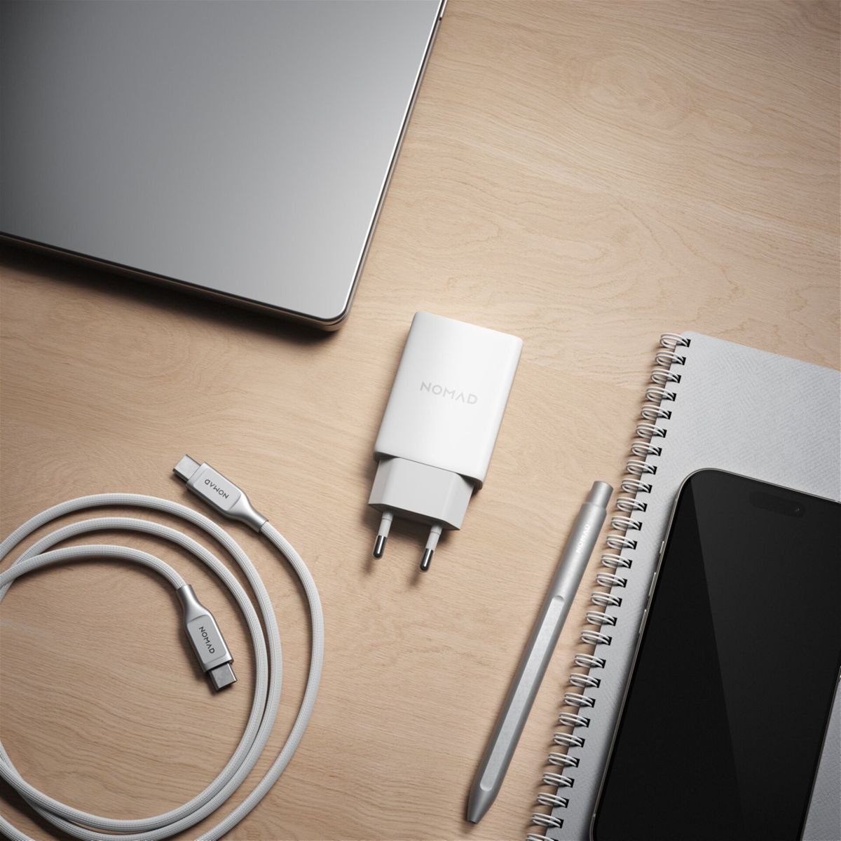 Ein weißes Nomad-Ladegerät, Kabel, Laptop, Telefon, Stift und Notizbuch auf einem Holztisch. Ladegerät und Kabel links.