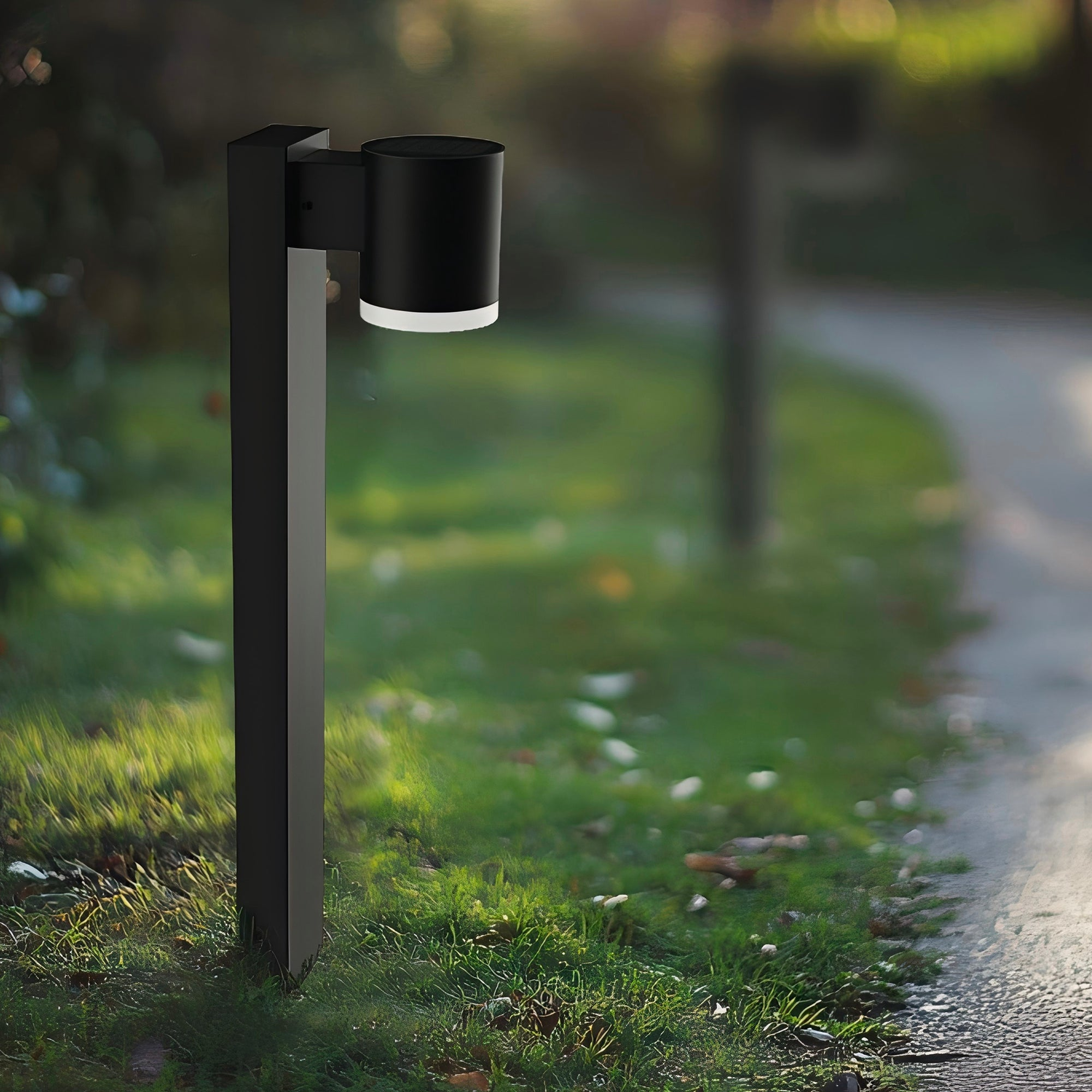 Eine schwarze Außenlaterne mit zylindrischem Kopf, im Gras neben einem Weg. Grüner und verschwommener Hintergrund.