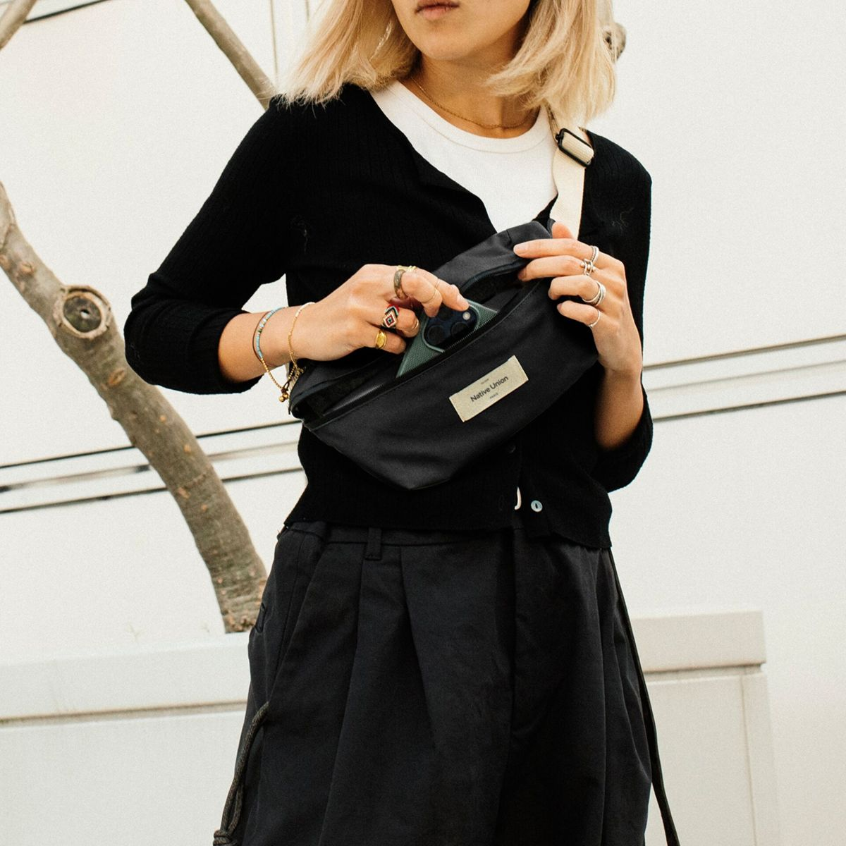 Eine Frau hält eine schwarze Bauchtasche und nimmt ein Telefon heraus. Sie trägt eine schwarze Strickjacke, schwarze Hose und Ringe. Der Hintergrund ist weiß.