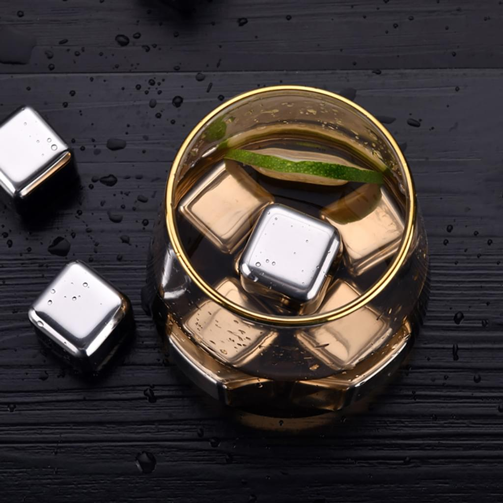 Ein Glas mit goldenen Metallwürfeln und einer Limettenscheibe auf einer schwarzen Holzoberfläche, umgeben von Metallwürfeln und Wassertropfen.