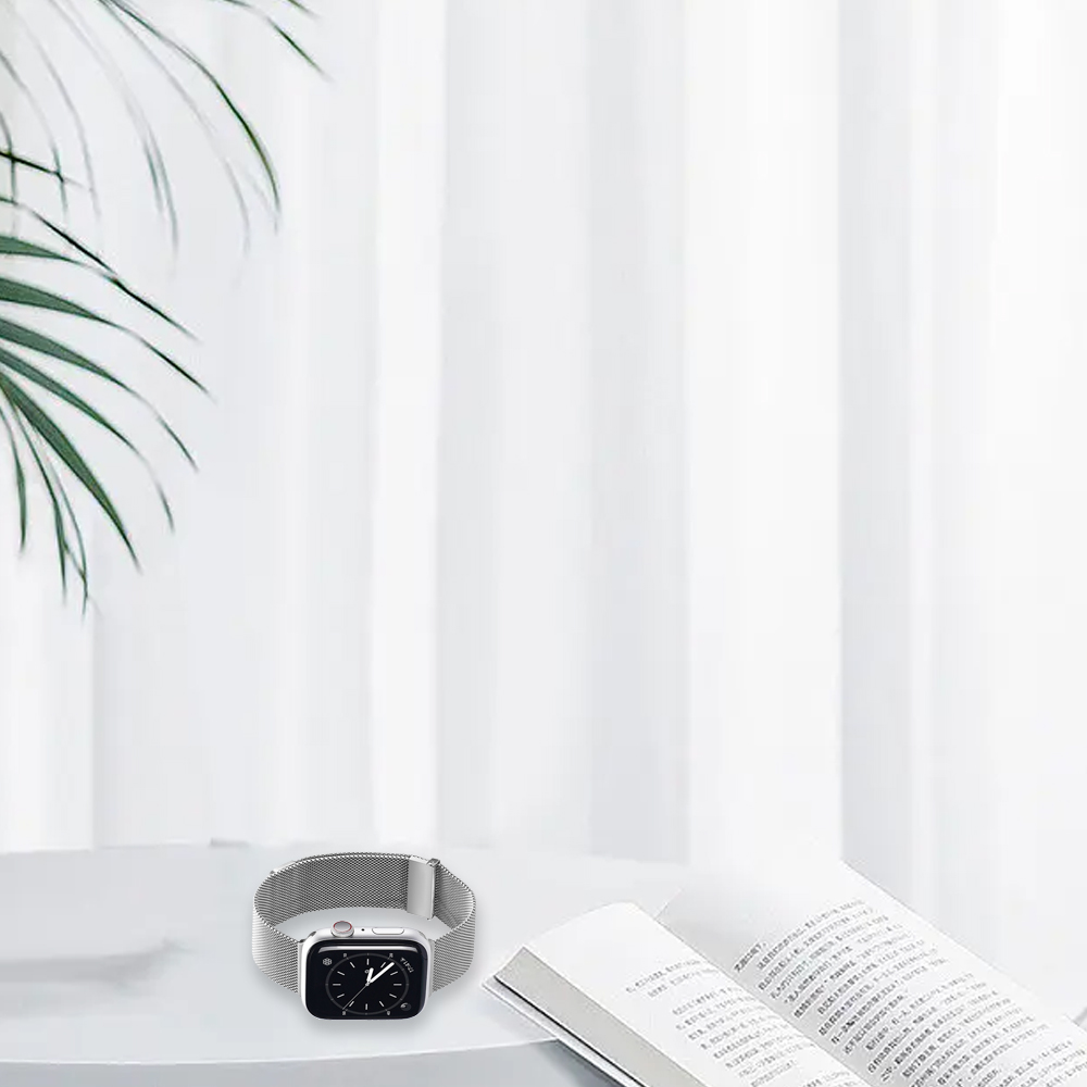 Eine silberne Smartwatch liegt auf einer weißen Oberfläche neben einem offenen Buch. Palmenblätter sind in der oberen linken Ecke. Weißer Vorhang im Hintergrund.