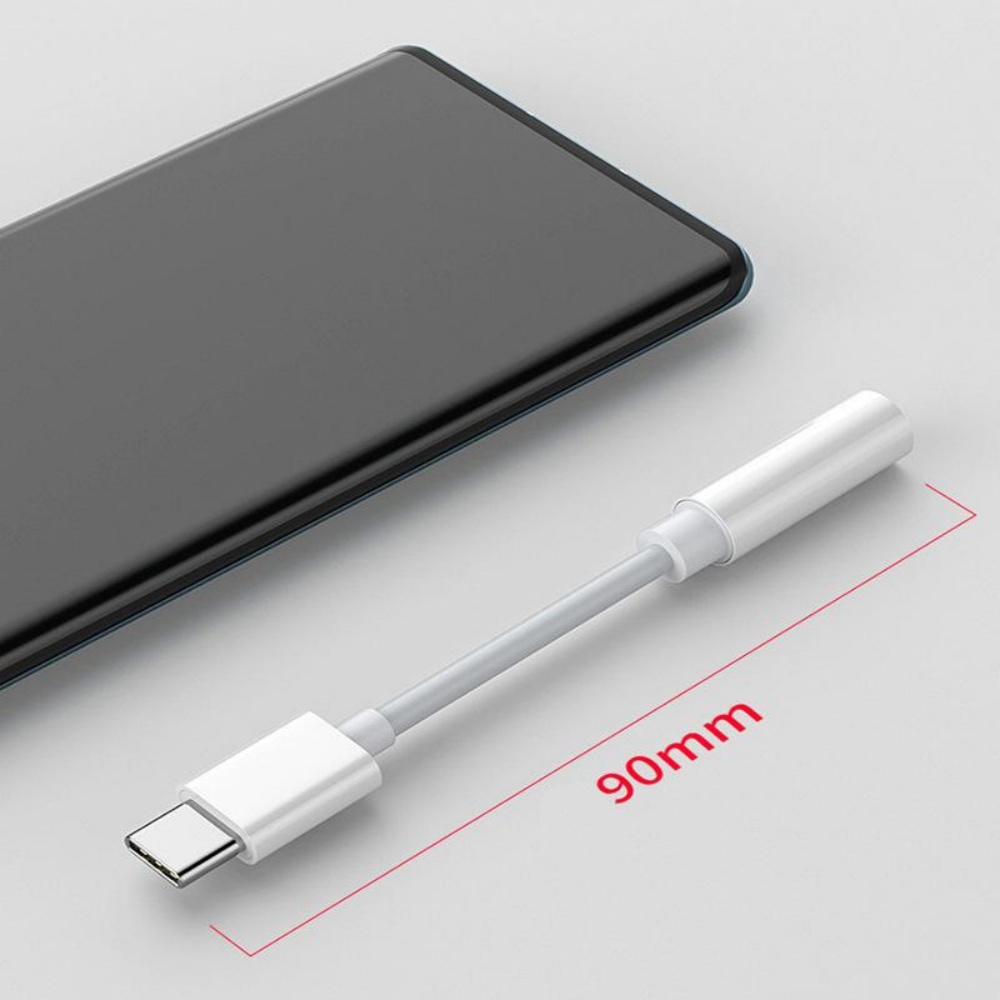 Ein weißer USB-C-auf-Kopfhöreranschlussadapter liegt in der Nähe eines schwarzen Smartphones. Der Adapter ist 90 mm lang. Weißer Hintergrund.