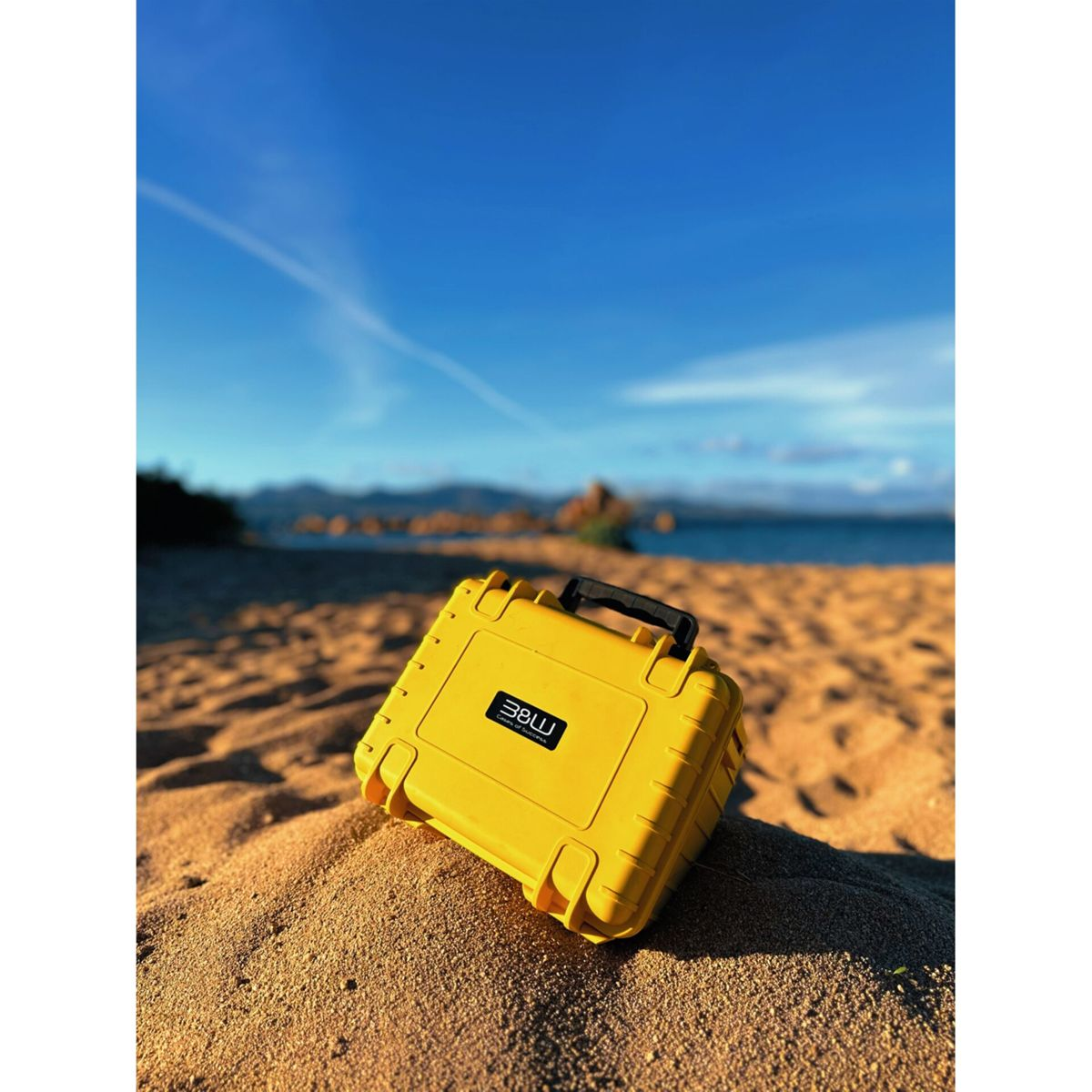 Ein gelber Koffer mit schwarzem Griff liegt auf Sand. Im Hintergrund sind Strand, Meer und blauer Himmel zu sehen.