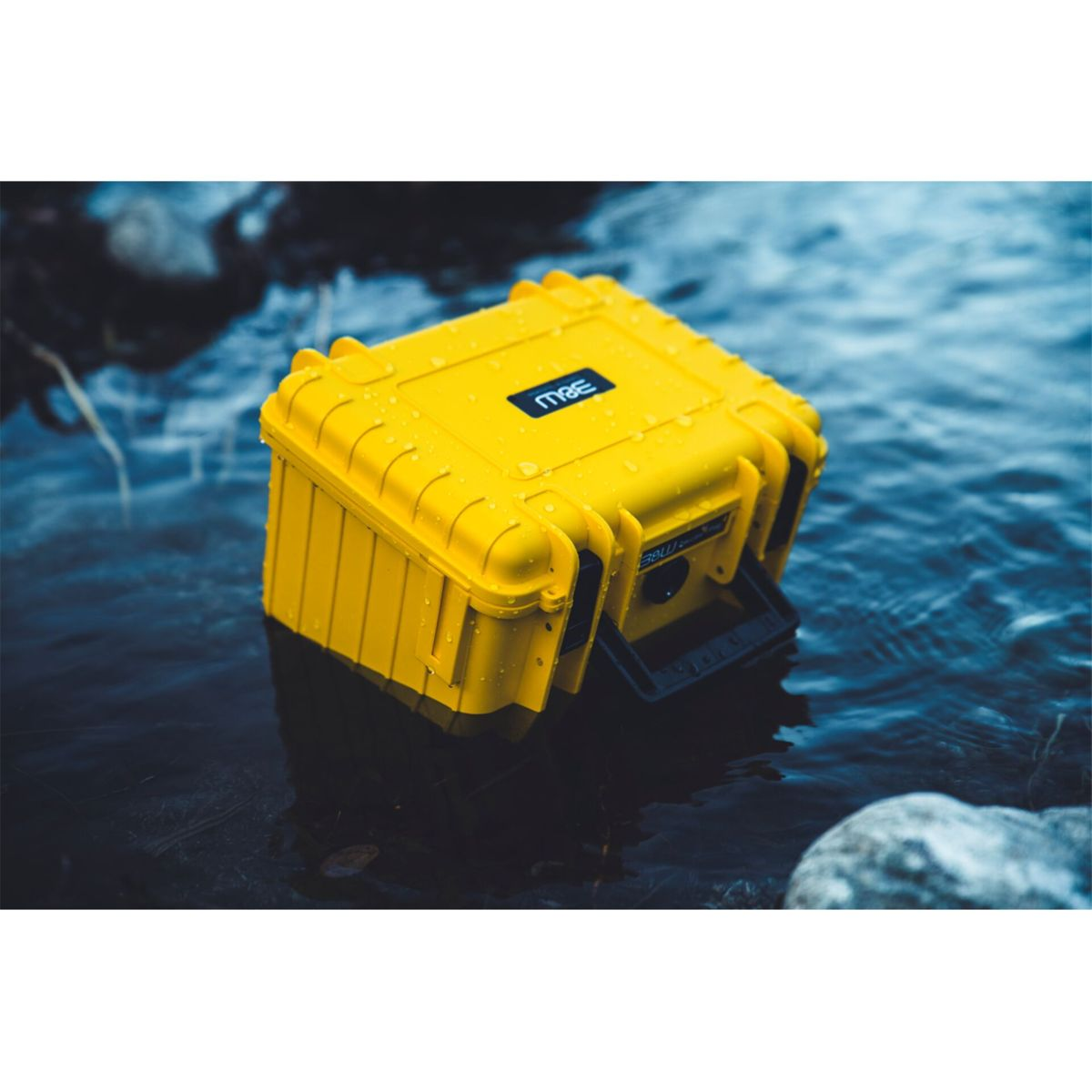 Eine gelbe, wasserdichte Box schwimmt im Wasser. Die Box hat einen schwarzen Griff und ein schwarzes Logo. Wassertropfen sind sichtbar.