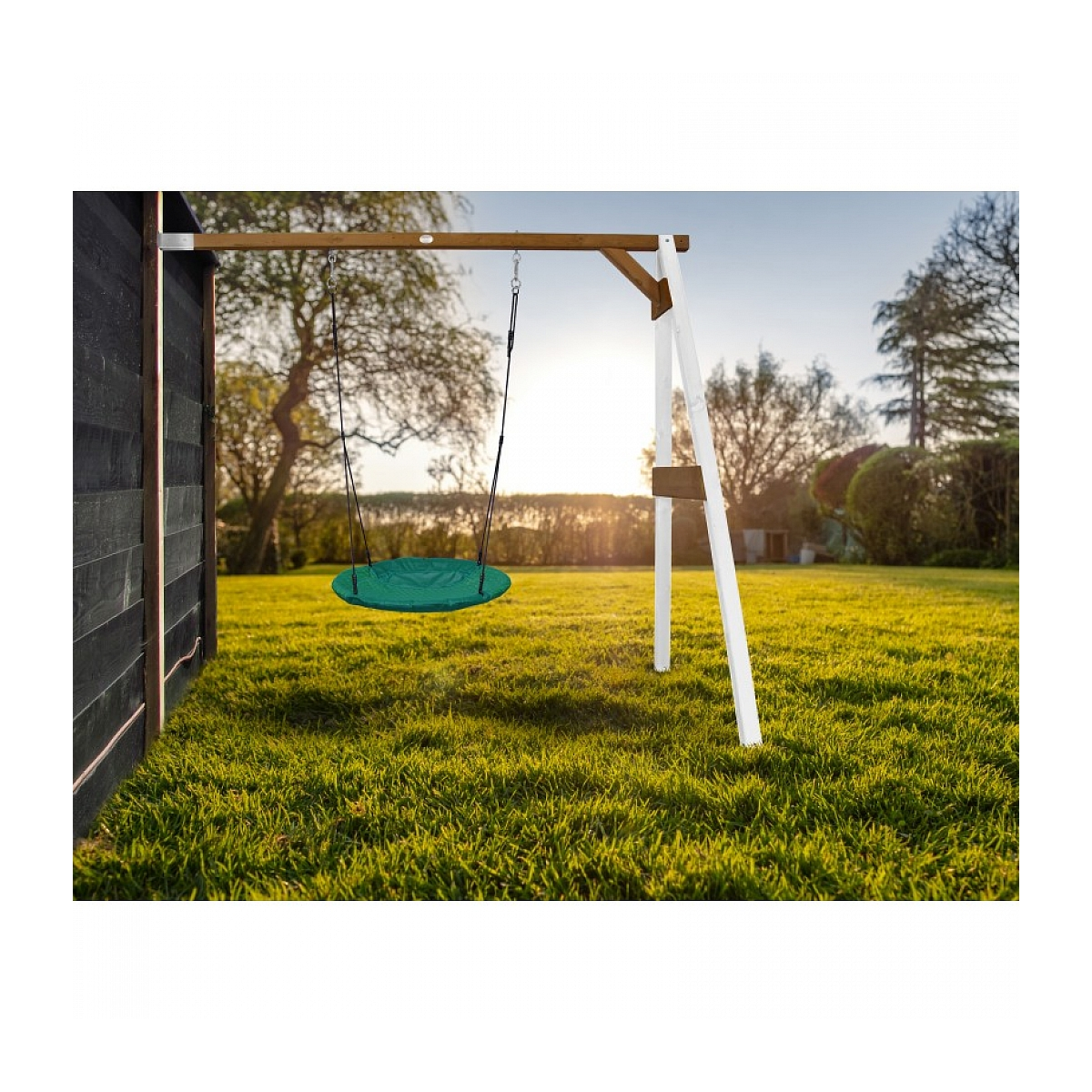 Eine grüne Schaukel hängt an einem Holzrahmen in einem sonnigen Garten. Gras bedeckt den Boden, und links ist ein schwarzer Zaun.