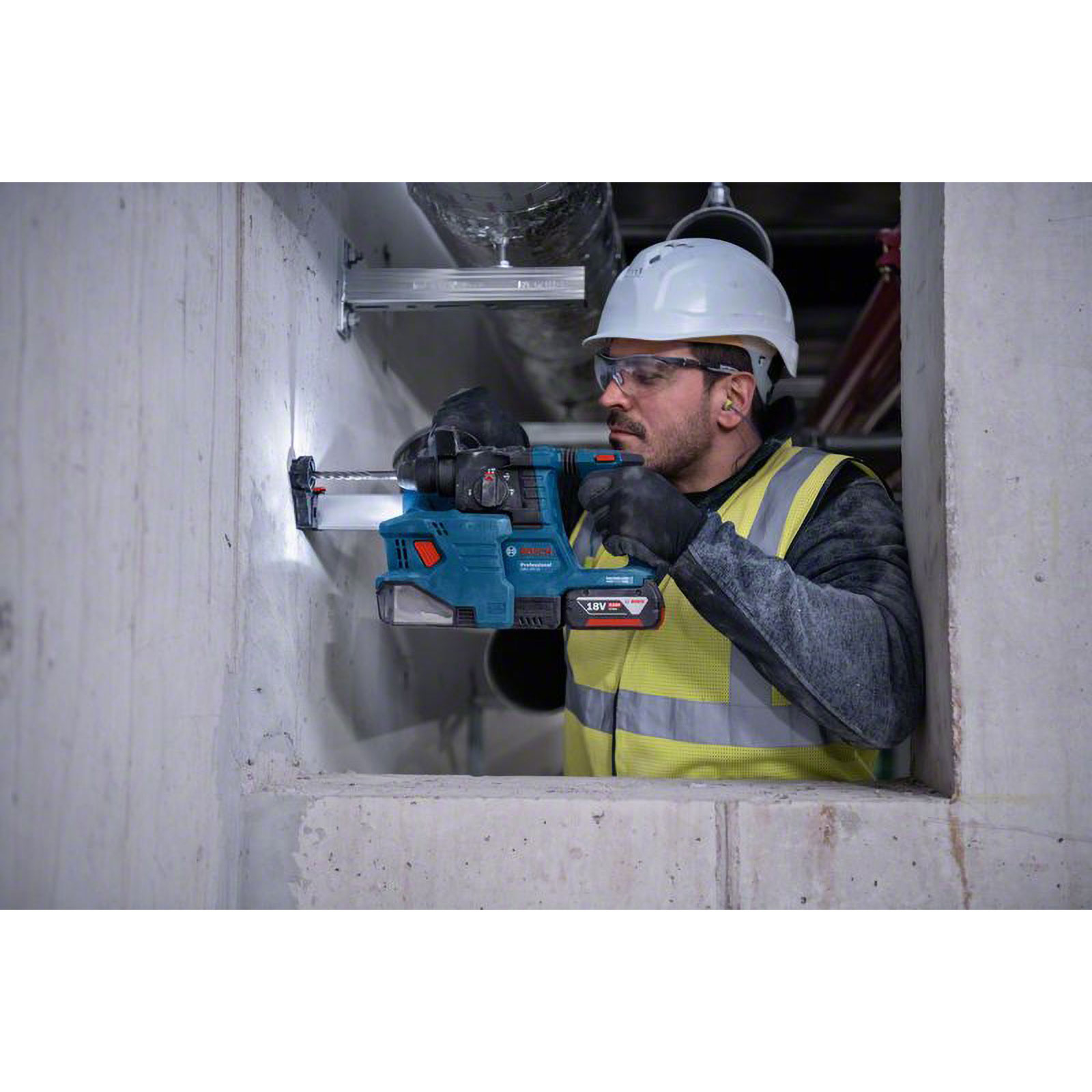 Ein Bauarbeiter bohrt in Beton. Er trägt einen Helm, eine Schutzbrille und eine Warnweste. Ein blauer Bohrer wird verwendet.