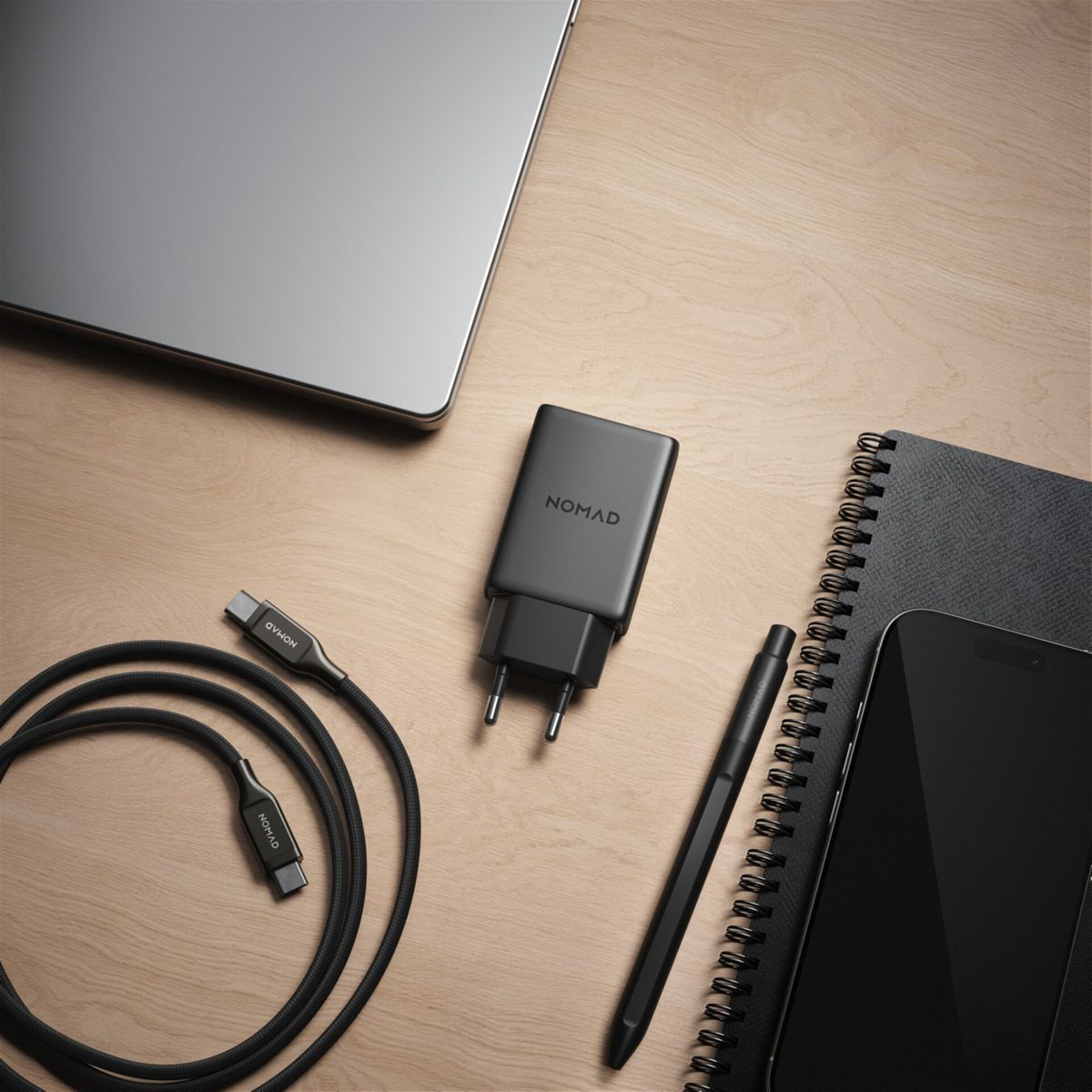 Ein Laptop, Ladegerät, Kabel, Stift, Telefon und Notizbuch liegen auf einer Holzoberfläche. Alle Gegenstände sind schwarz.