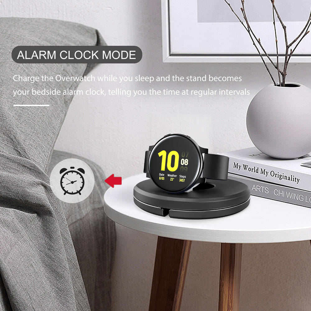 Eine Smartwatch auf einer Ladestation zeigt die Uhrzeit an. Ein Wecker-Symbol und der Text 'ALARM CLOCK MODE' sind sichtbar. Ein weißer Tisch ist im Bild.