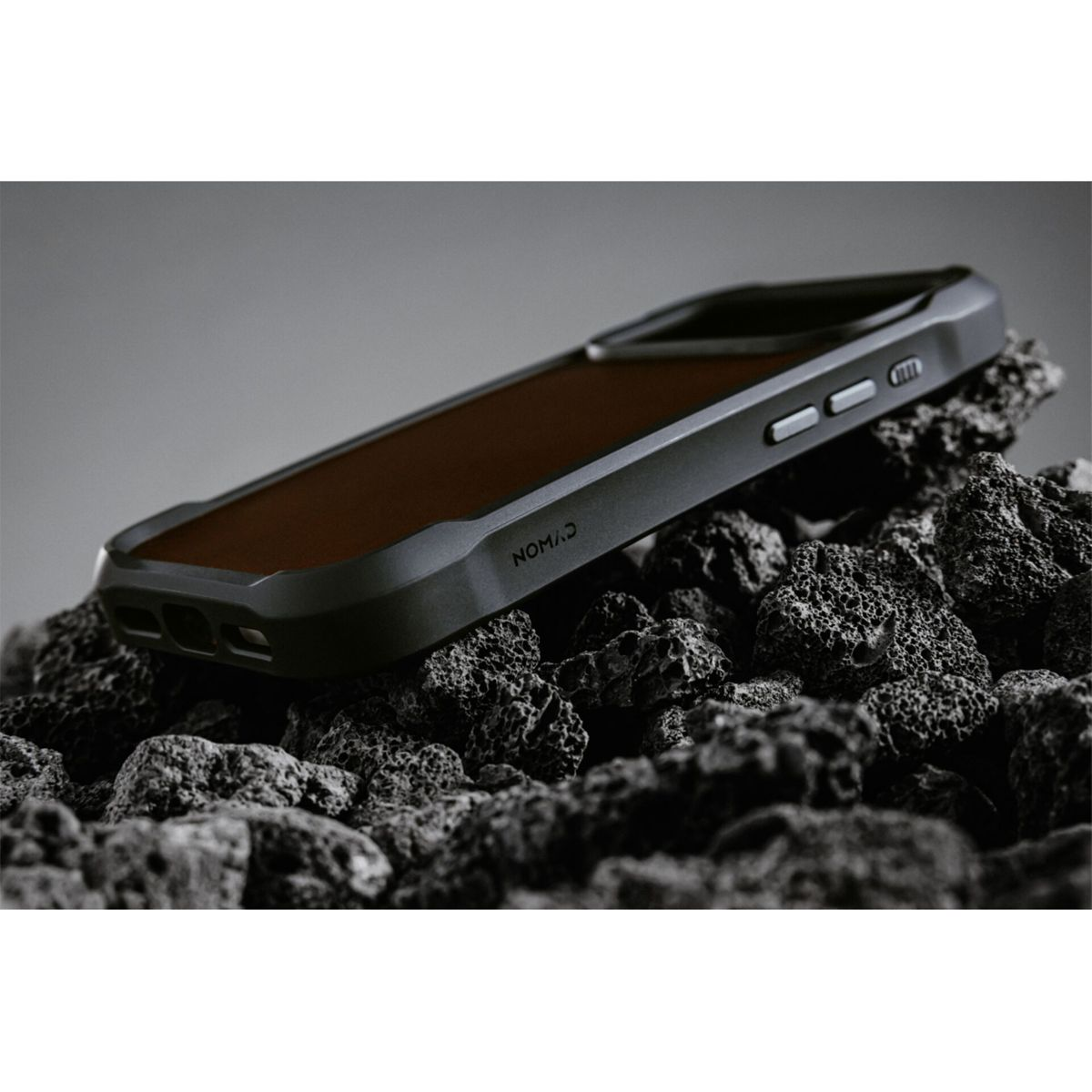 Una custodia per telefono con retro in pelle marrone su rocce nere. La custodia ha bordi grigi e il marchio 'NOMAD'.