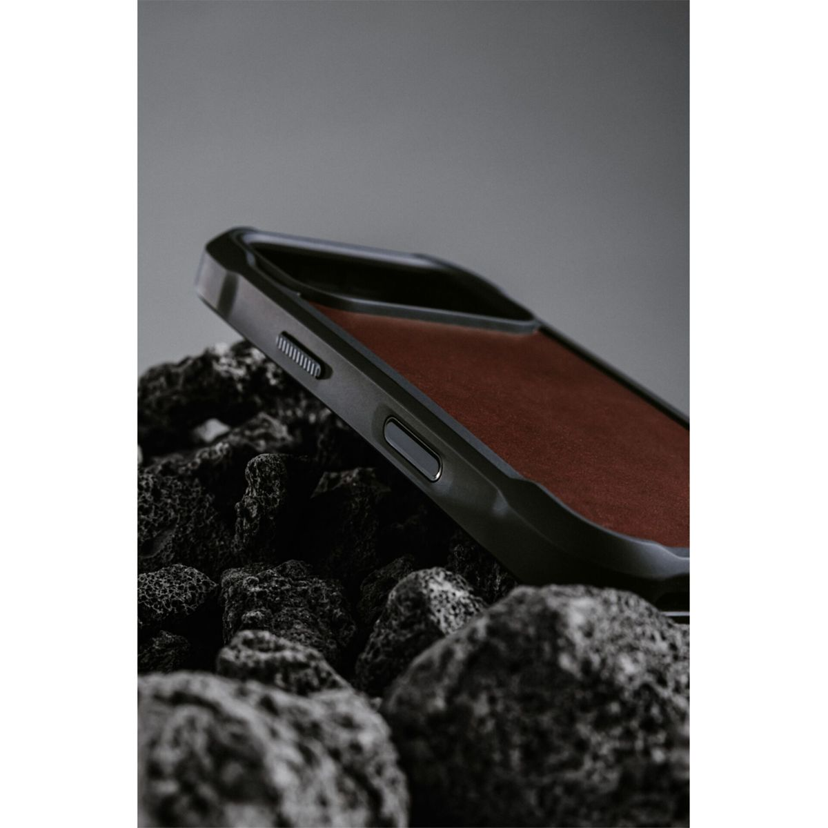 Un telefono con custodia nera poggia su rocce scure e strutturate. Lo schermo del telefono è marrone scuro, lo sfondo è grigio.