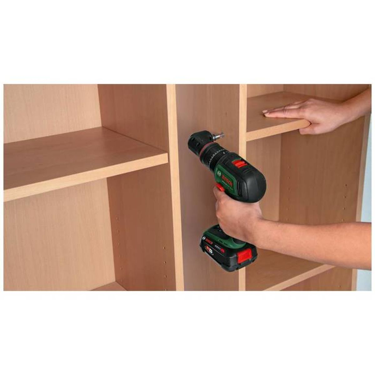 Eine Person verwendet einen schwarz-grünen Akkuschrauber, um einen Holzschrank zusammenzubauen. Der Schrank hat leere Regale.