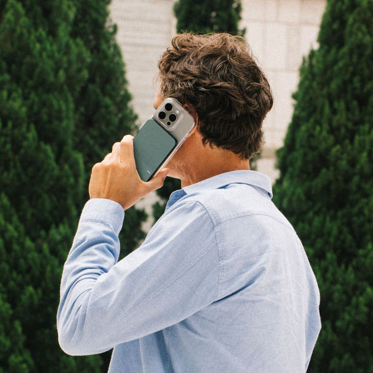 Ein Mann mit braunen Haaren hält ein Telefon an sein Ohr. Er trägt ein blaues Hemd und steht vor grünen Bäumen.