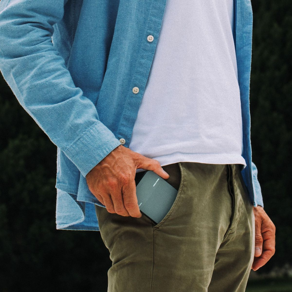 Ein Mann in blauem Hemd und grüner Hose steckt einen grauen Lautsprecher in seine Tasche. Ein weißes T-Shirt ist sichtbar.