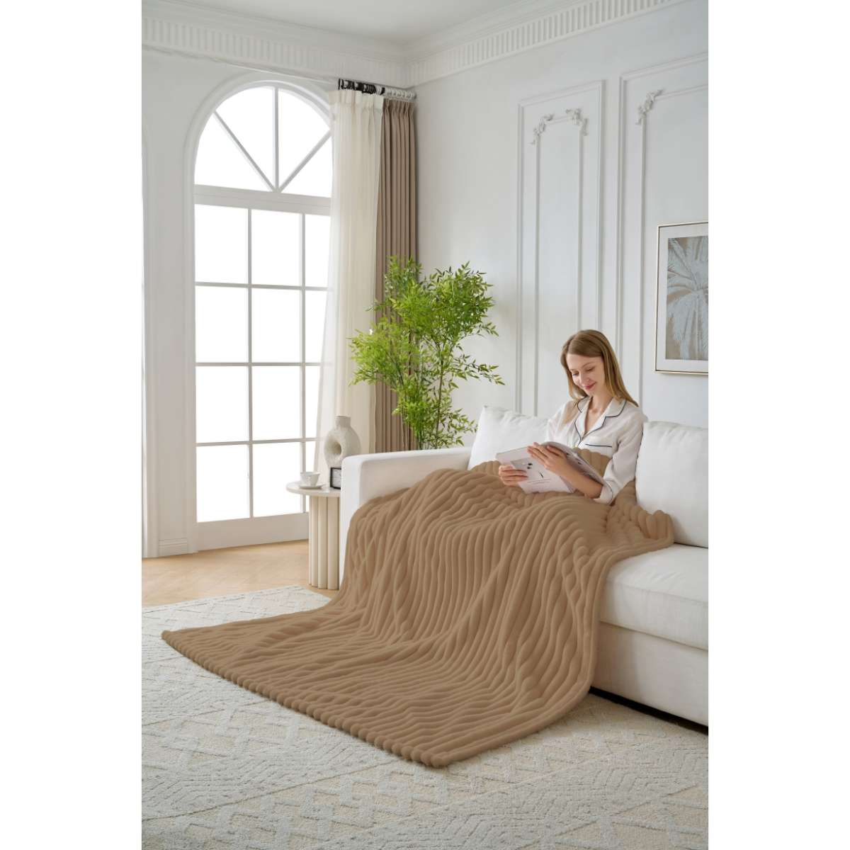 Eine Frau sitzt auf einem weißen Sofa und liest unter einer braunen Decke. Im Hintergrund sind ein Fenster und eine Pflanze zu sehen. Der Teppich ist beige.