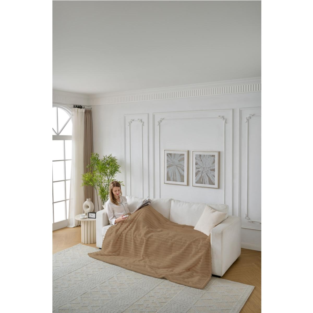 Frau liest unter einer braunen Decke auf einem weißen Sofa. Der Raum hat weiße Wände, einen Teppich, ein Fenster und dekorative Kunst.