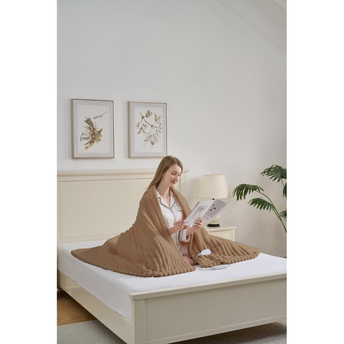 Eine Frau sitzt auf einem Bett, bedeckt von einer braunen Decke, und liest eine Zeitschrift. Zwei gerahmte Bilder hängen an der Wand, eine Lampe steht auf dem Nachttisch.