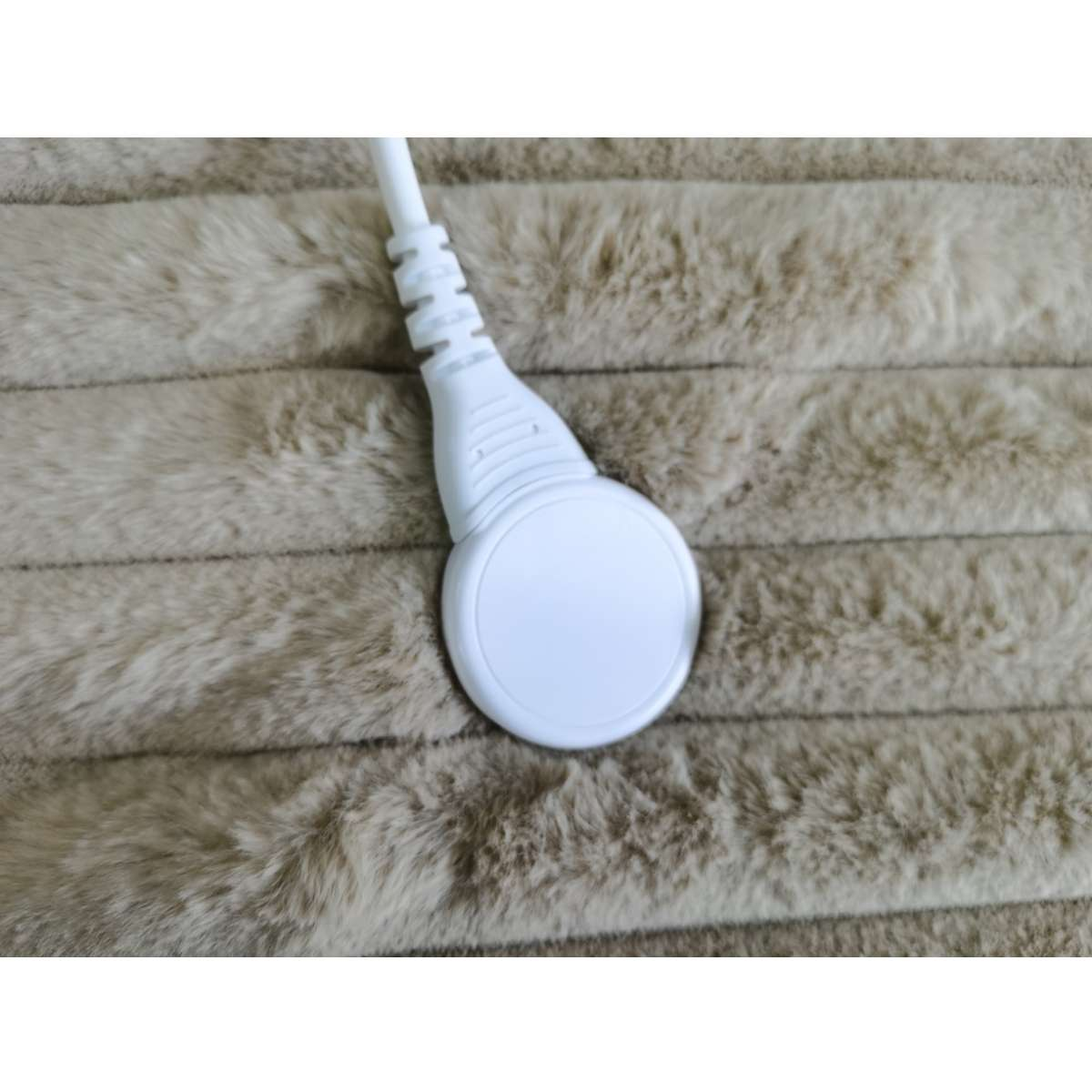 Ein weißes, rundes Gerät mit Netzkabel liegt auf einer beigen, flauschigen Oberfläche. Das Gerät ist mittig.