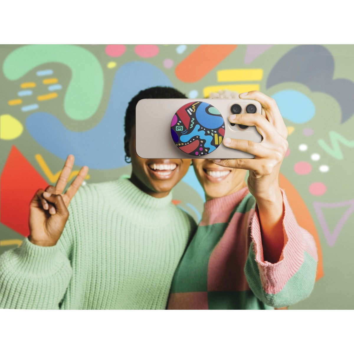 Due donne sorridenti scattano un selfie con un telefono. Il telefono ha un PopSocket colorato. Lo sfondo è un muro verde con forme.