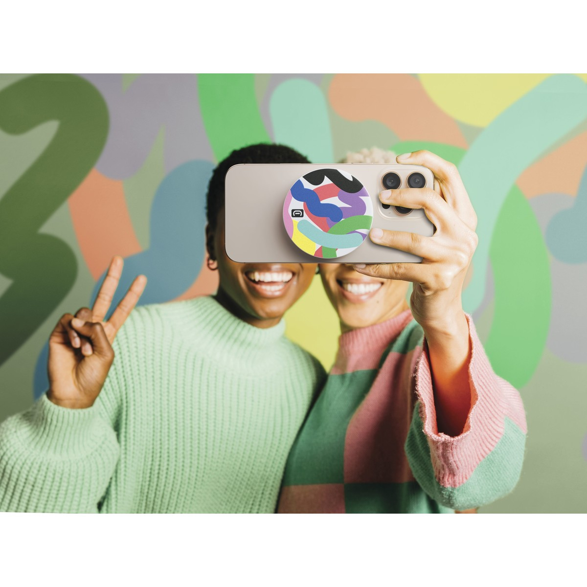 Due donne sorridenti si fanno un selfie con un telefono. Una mostra il segno della pace. Lo sfondo è un motivo astratto e colorato.