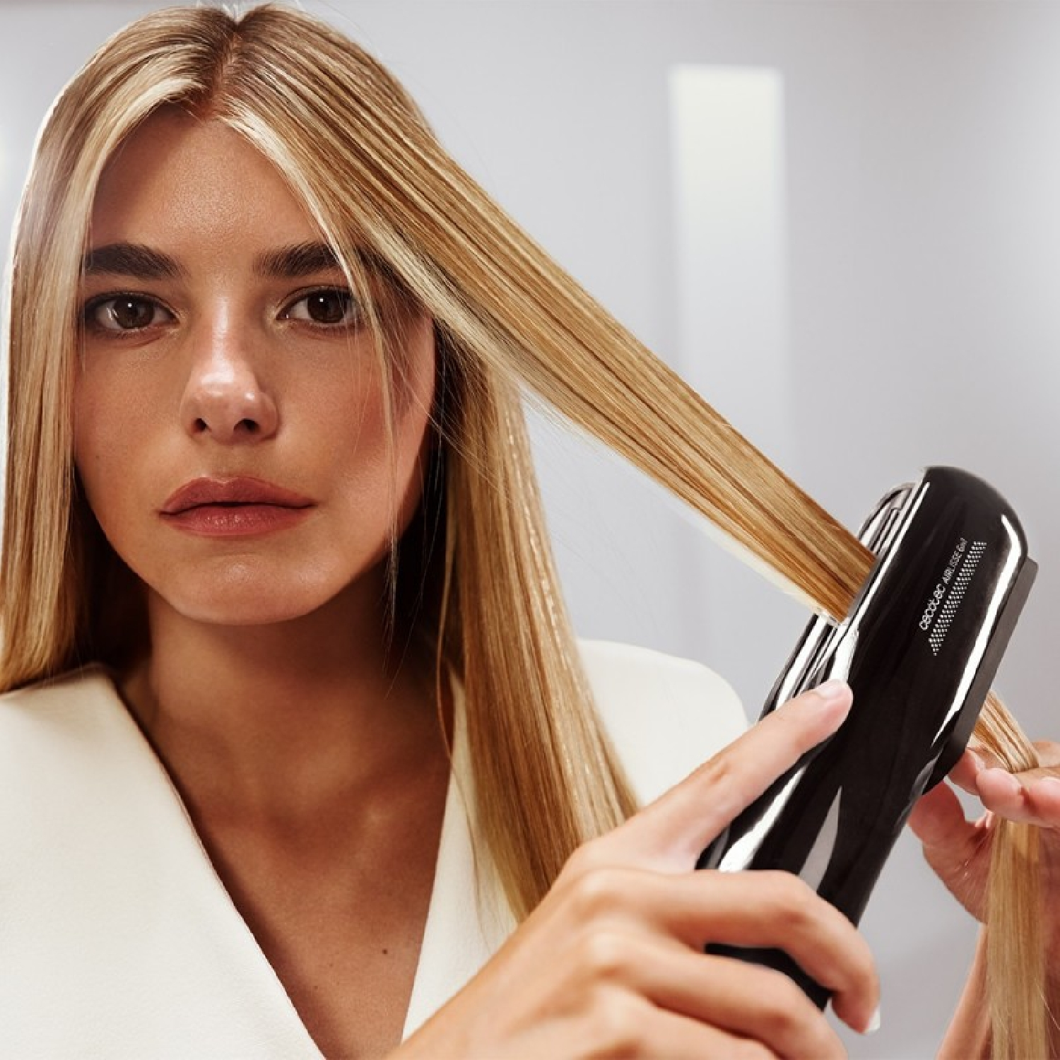 Una donna con i capelli biondi usa una piastra nera. Guarda la fotocamera. Lo sfondo è bianco e grigio.