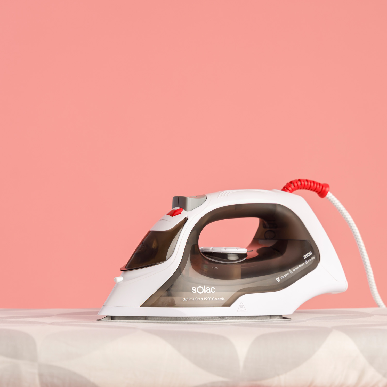 Ein weißes und schwarzes Bügeleisen mit rotem Griff und Kabel liegt auf einem gemusterten Bügelbrett. Der Hintergrund ist eine rosa Wand.