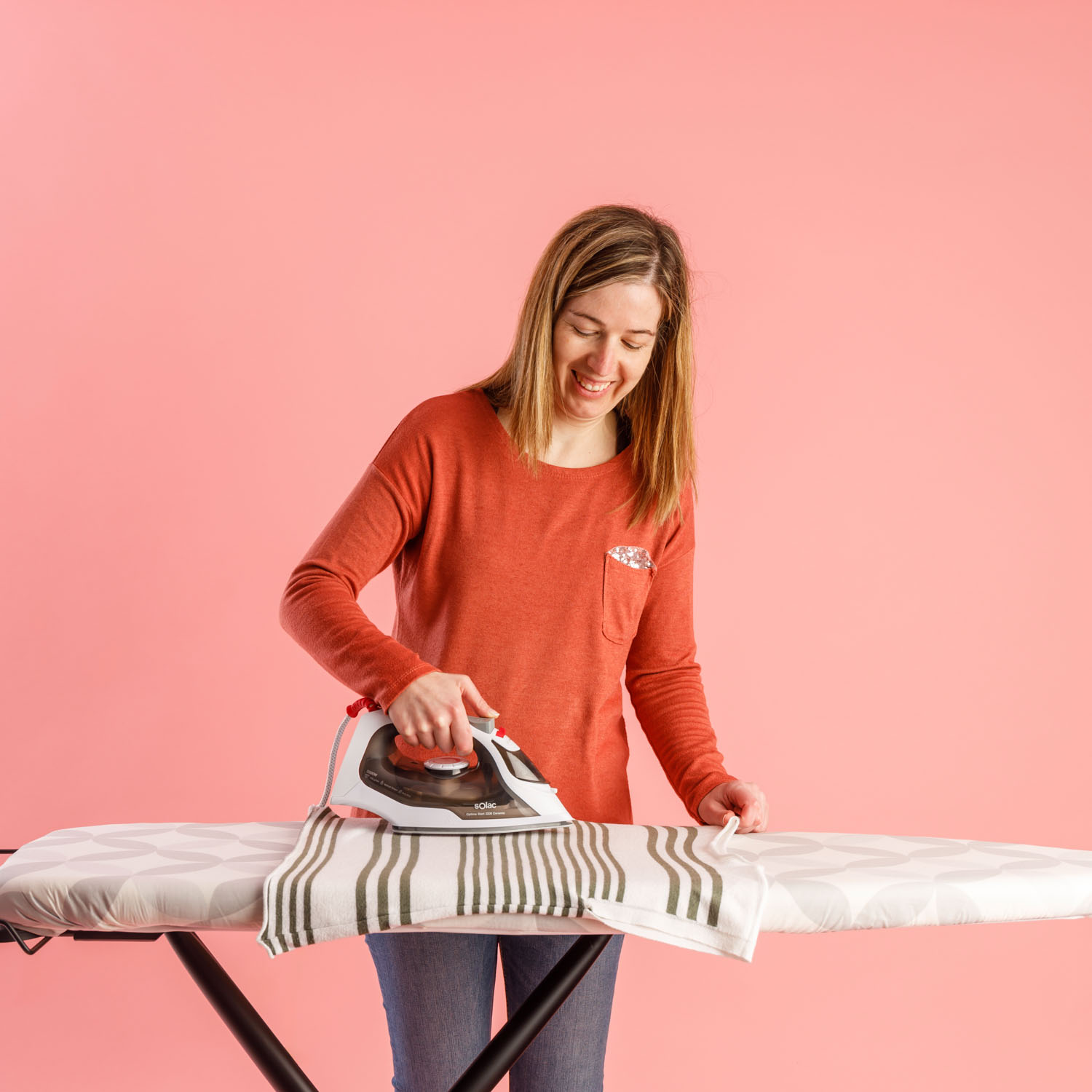 Eine Frau bügelt ein gestreiftes Handtuch auf einem Bügelbrett vor rosa Hintergrund. Sie trägt ein rotes Hemd und Jeans und lächelt.