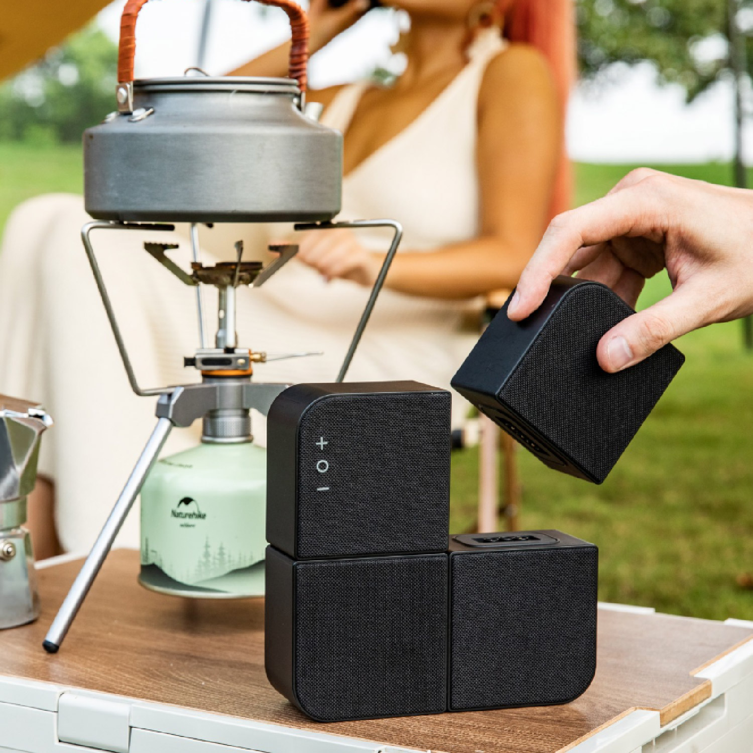 Una escena de camping con altavoces negros que se están colocando. Una persona está sosteniendo uno. También hay una tetera y una estufa en la mesa.