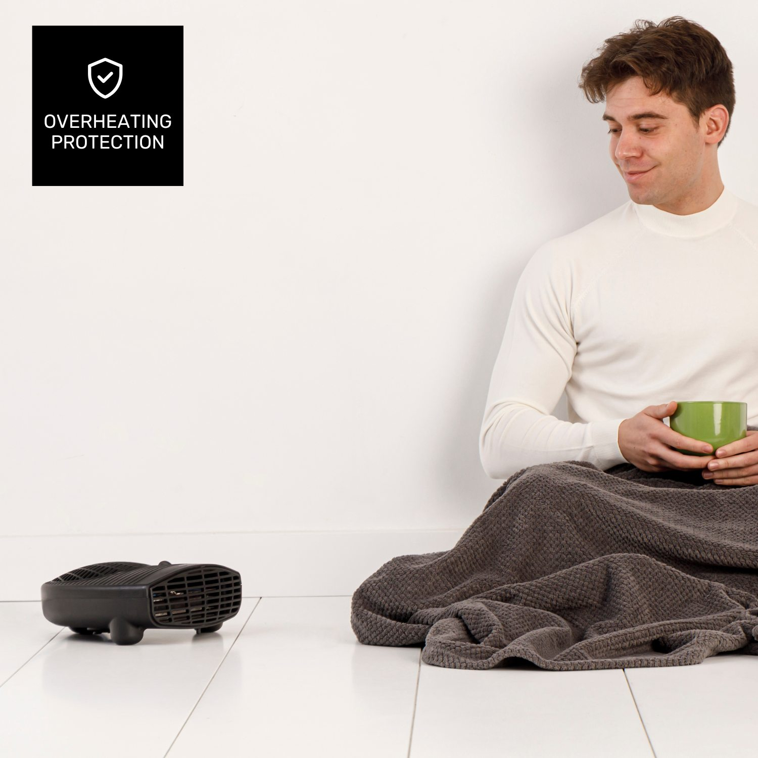 Hombre con taza sentado cerca de un calefactor, cubierto con una manta. Etiqueta de protección contra sobrecalentamiento. Fondo y piso blancos.