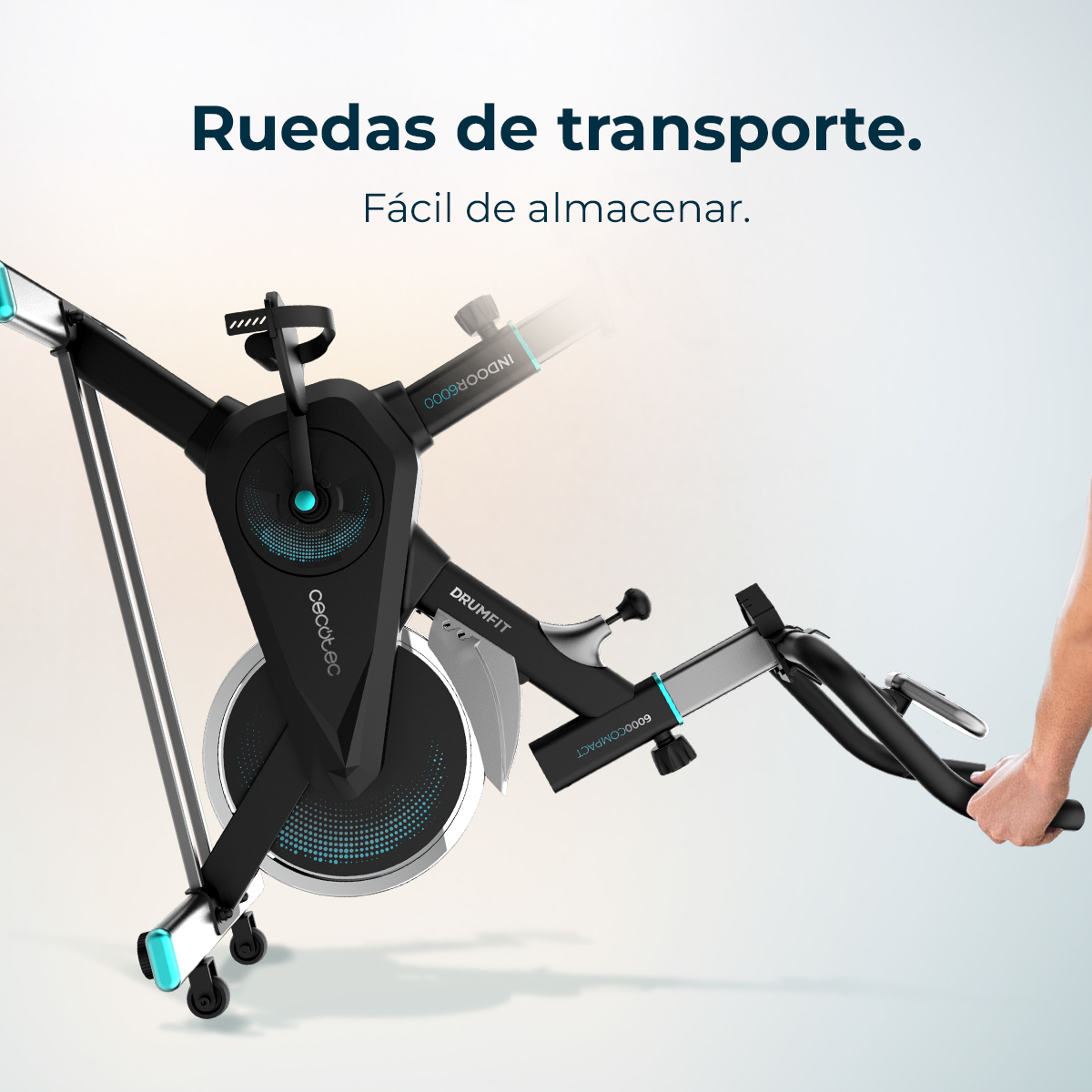 Bicicleta estática negra con detalles plateados. Una mano sujeta el manillar. La bicicleta tiene ruedas. Texto superior: 'Ruedas de transporte'.